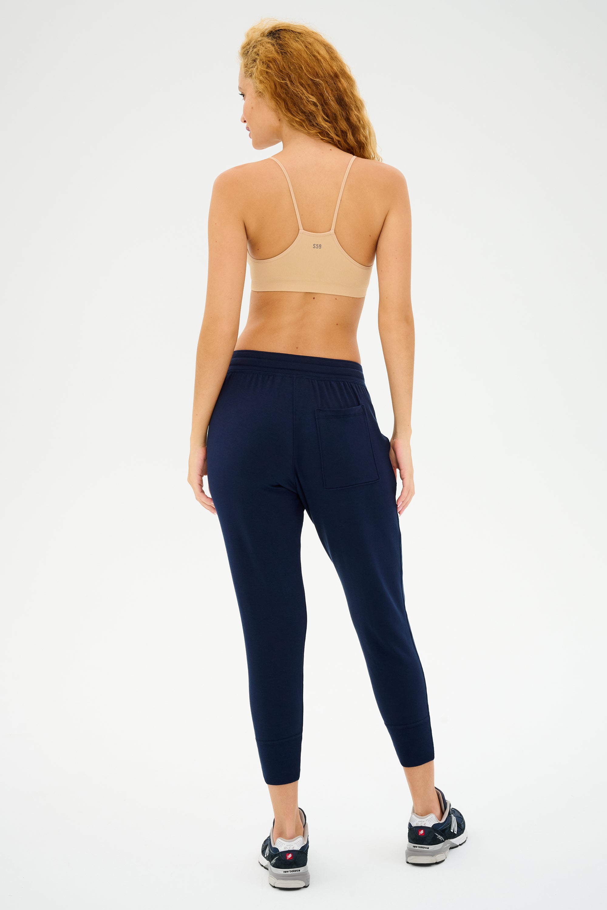 A person with curly hair, wearing a Splits59 Loren Seamless Bra in Nude and navy joggers, standing turned away.