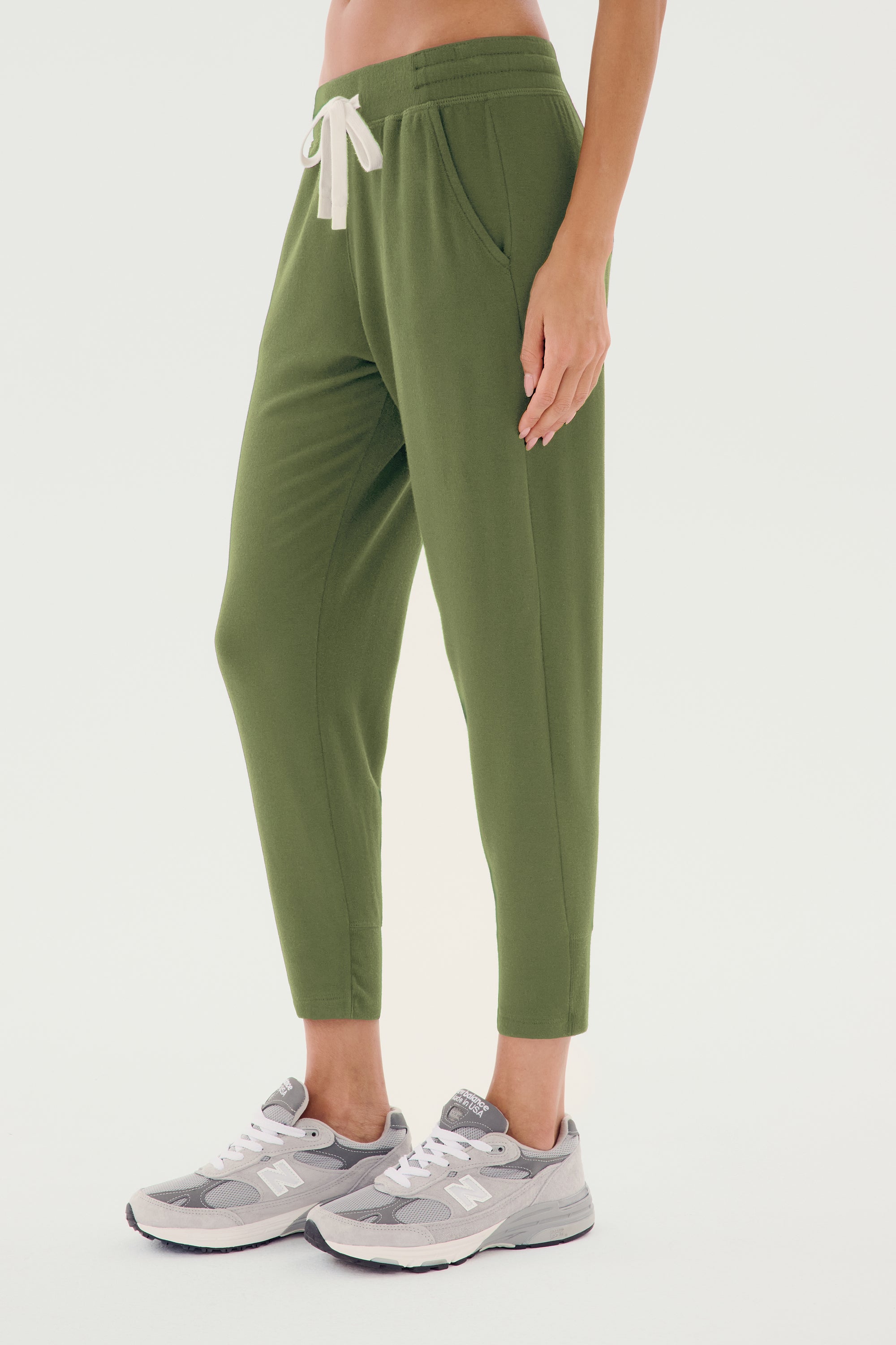 A person wearing SPLITS59's Reena Fleece 7/8 Sweatpant in Olive, featuring front pockets and a white drawstring, paired with gray sneakers and complemented by a classic rugby polo, stands against a plain background.