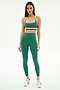 A woman poses in a studio wearing the SPLITS59 Bianca High Waist Rigor 7/8 leggings and matching sports bra in Arugula/White, crafted from soft nylon spandex fabric, paired with white sneakers against a white background.