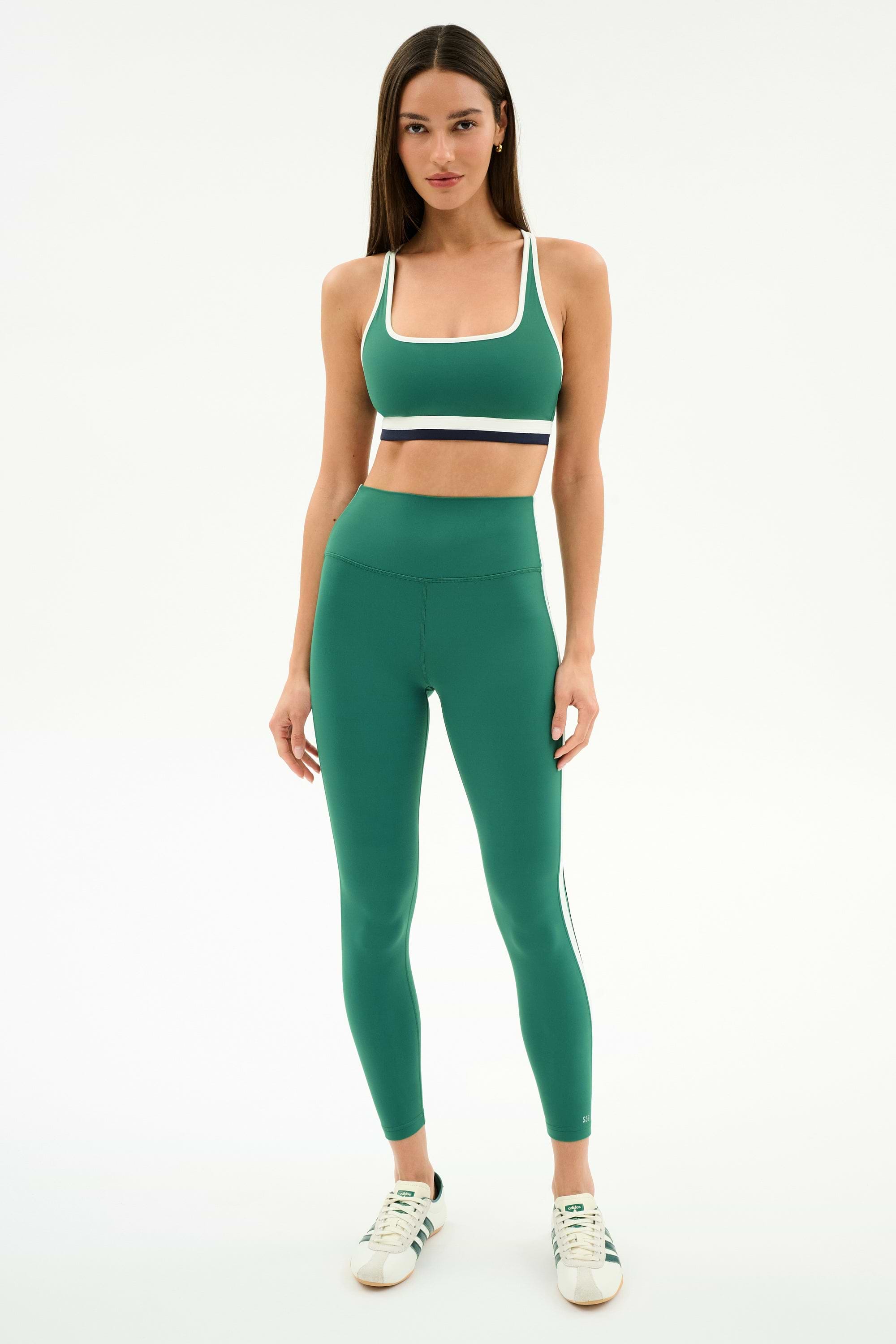 A woman stands against a white background, wearing the SPLITS59 Bianca Rigor Bra in Arugula/White with matching leggings in a soft nylon spandex blend, paired with white sneakers.