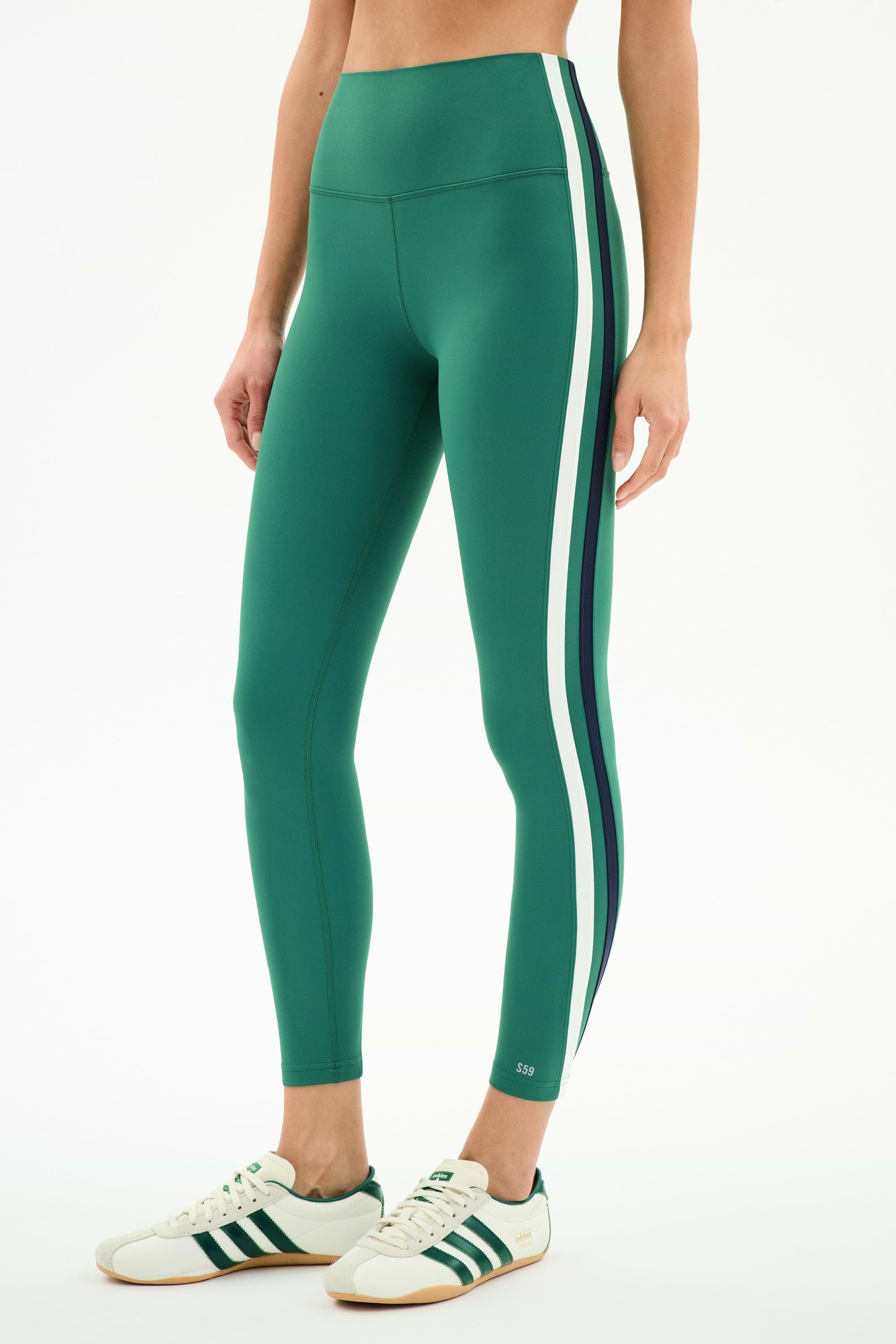 A person stands against a plain background wearing SPLITS59 Bianca High Waist Rigor 7/8 leggings in Arugula/White, which feature white and dark green side stripes, paired with white sneakers with green stripes.