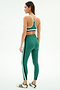 A woman stands at a slight angle against a plain background, wearing SPLITS59's Bianca High Waist Rigor 7/8 leggings in Arugula/White with white and navy side stripes, paired with a white-trimmed sports bra and white sneakers.