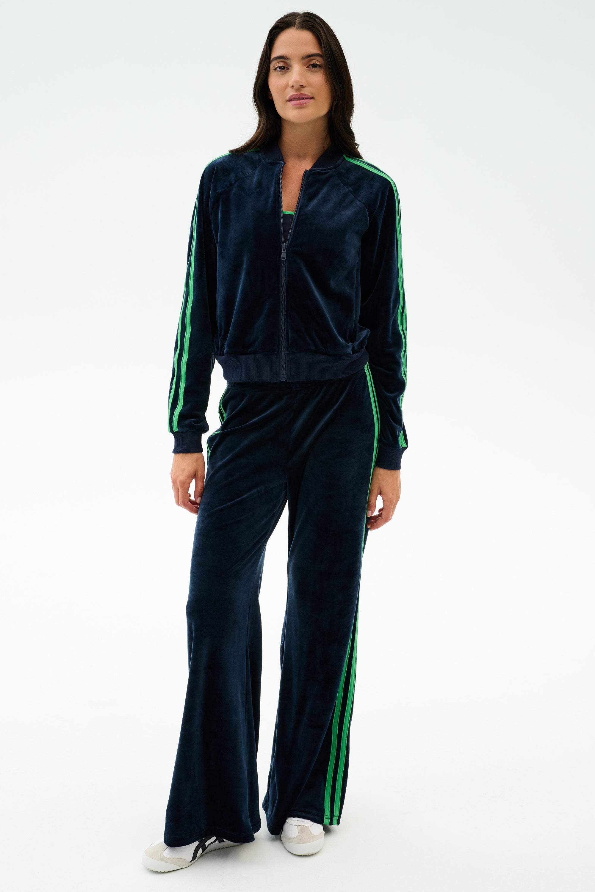 A woman models the SPLITS59 Heritage Velour Bomber in Indigo/Grass, featuring green stripes, styled with white sneakers—ideal for yoga or lounging—against a plain light background.