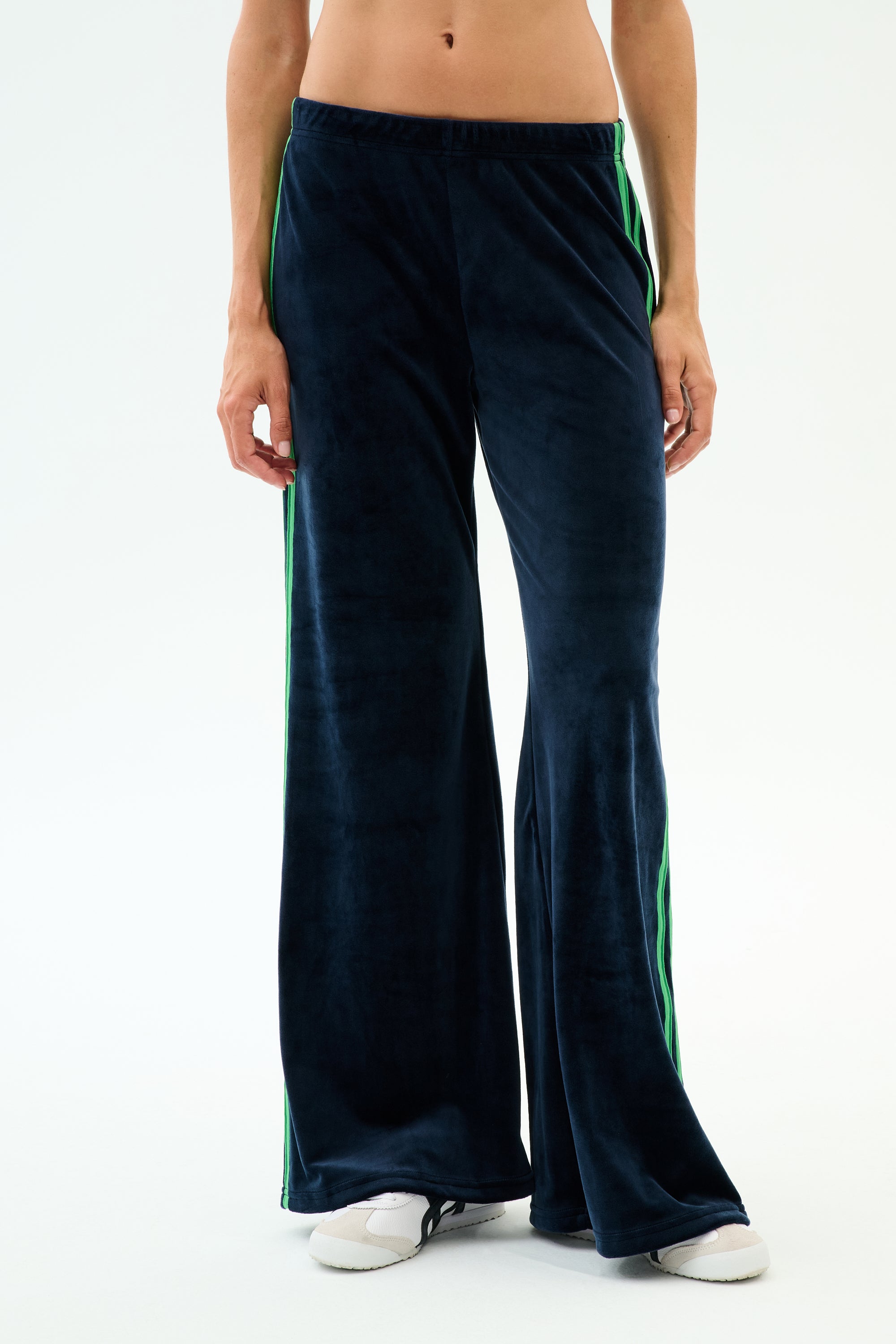 A person models the SPLITS59 Heritage Velour Pant in Indigo/Grass, featuring wide-leg navy blue velour fabric with green side stripes, ankles visible above white sneakers against a plain white background.