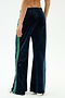 A person is seen from behind wearing the SPLITS59 Heritage Velour Pant in Indigo/Grass, featuring navy velour fabric with green side stripes and a relaxed wide-leg fit that flares stylishly at the ankle, against a plain white background.