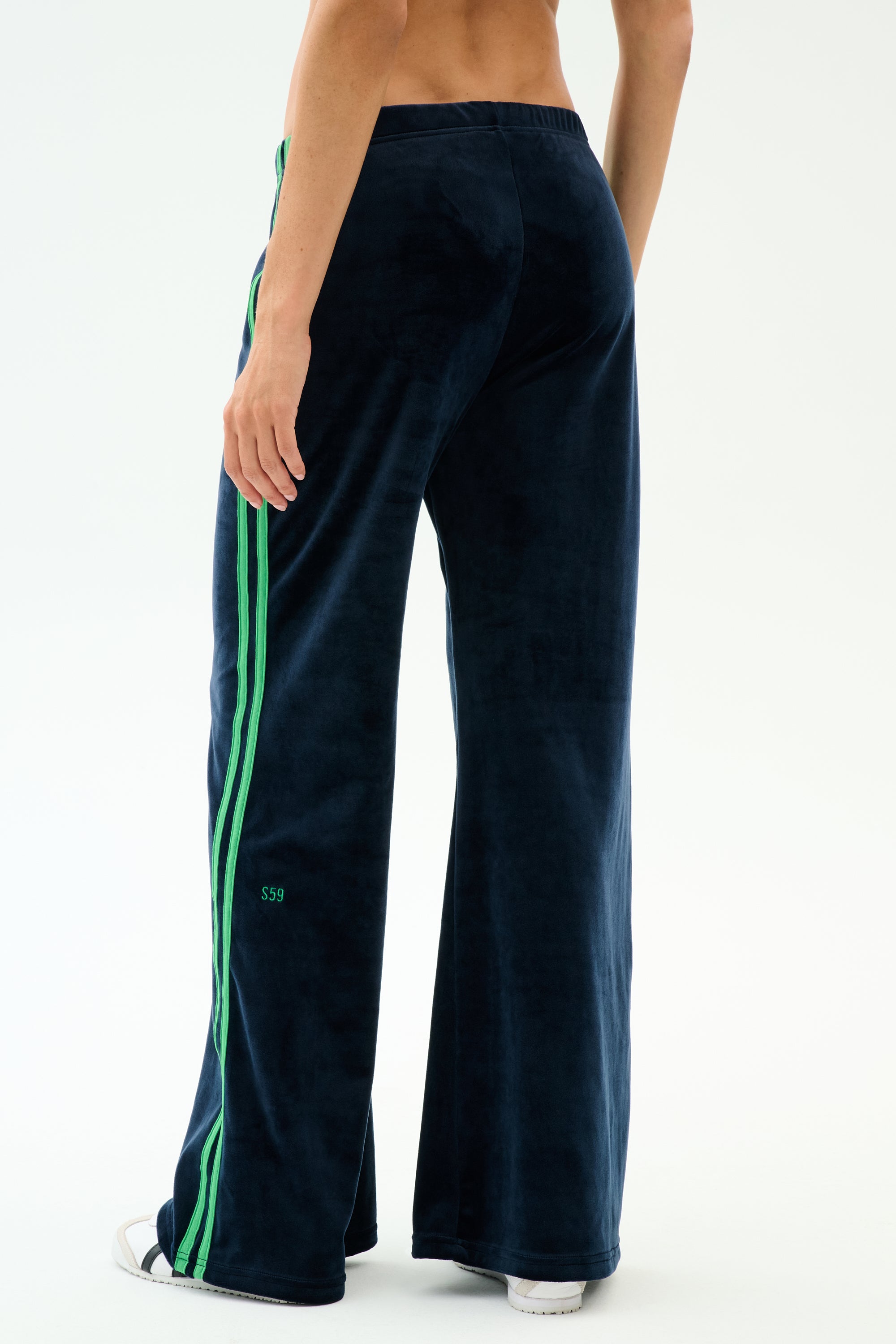 A person is seen from behind wearing the SPLITS59 Heritage Velour Pant in Indigo/Grass, featuring navy velour fabric with green side stripes and a relaxed wide-leg fit that flares stylishly at the ankle, against a plain white background.