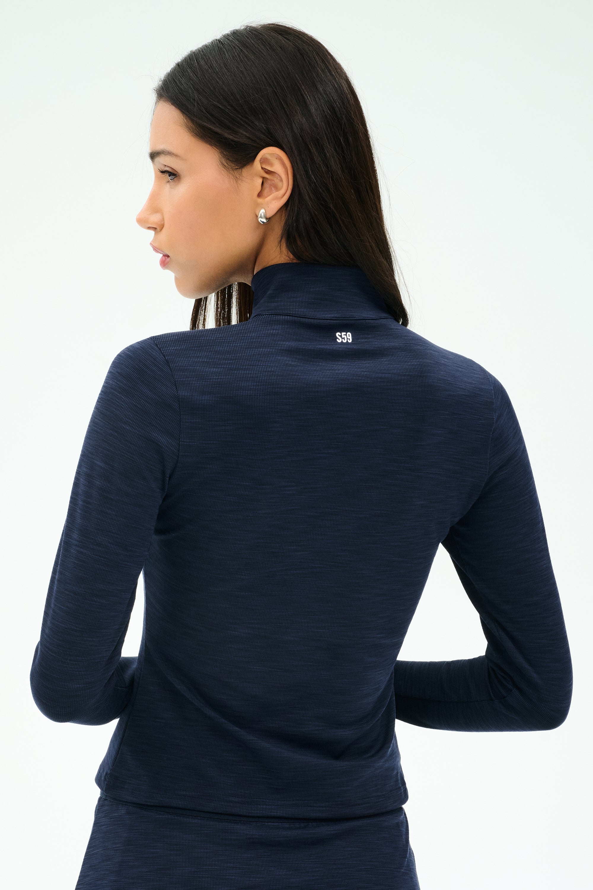 A woman with long dark hair is seen from behind, wearing the SPLITS59 Stevie Active Rib Half Zip Waist Length in Heather Indigo, a polyester spandex top with "SSG" on the back collar, against a plain light background.