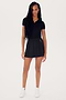 A woman in a black polo shirt and SPLITS59's Venus High Waist Rigor Skort w/Piping - Black stands facing the camera, wearing white sneakers, with long braided hair.