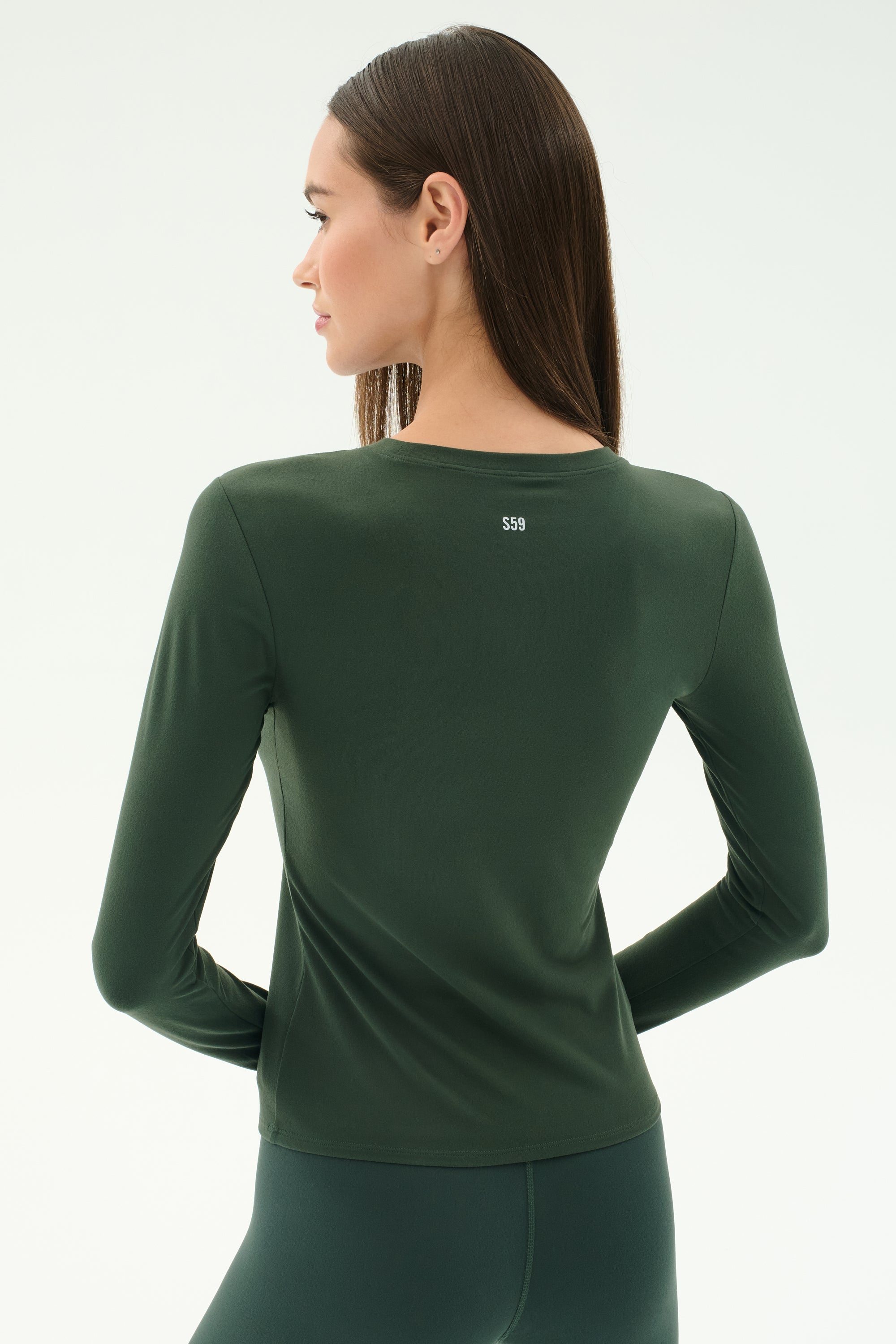 A woman with straight brown hair models the SPLITS59 Airweight Lite LS Waist Length - Military fitted top, paired with matching spandex bottoms, facing away from the camera against a plain light background.