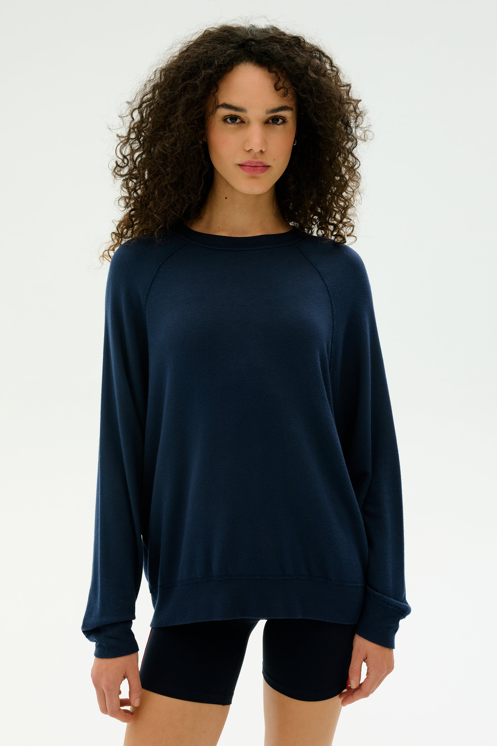 A woman with curly hair wears the SPLITS59 Andie Fleece Sweatshirt in indigo, paired with matching shorts, standing against a plain white background.
