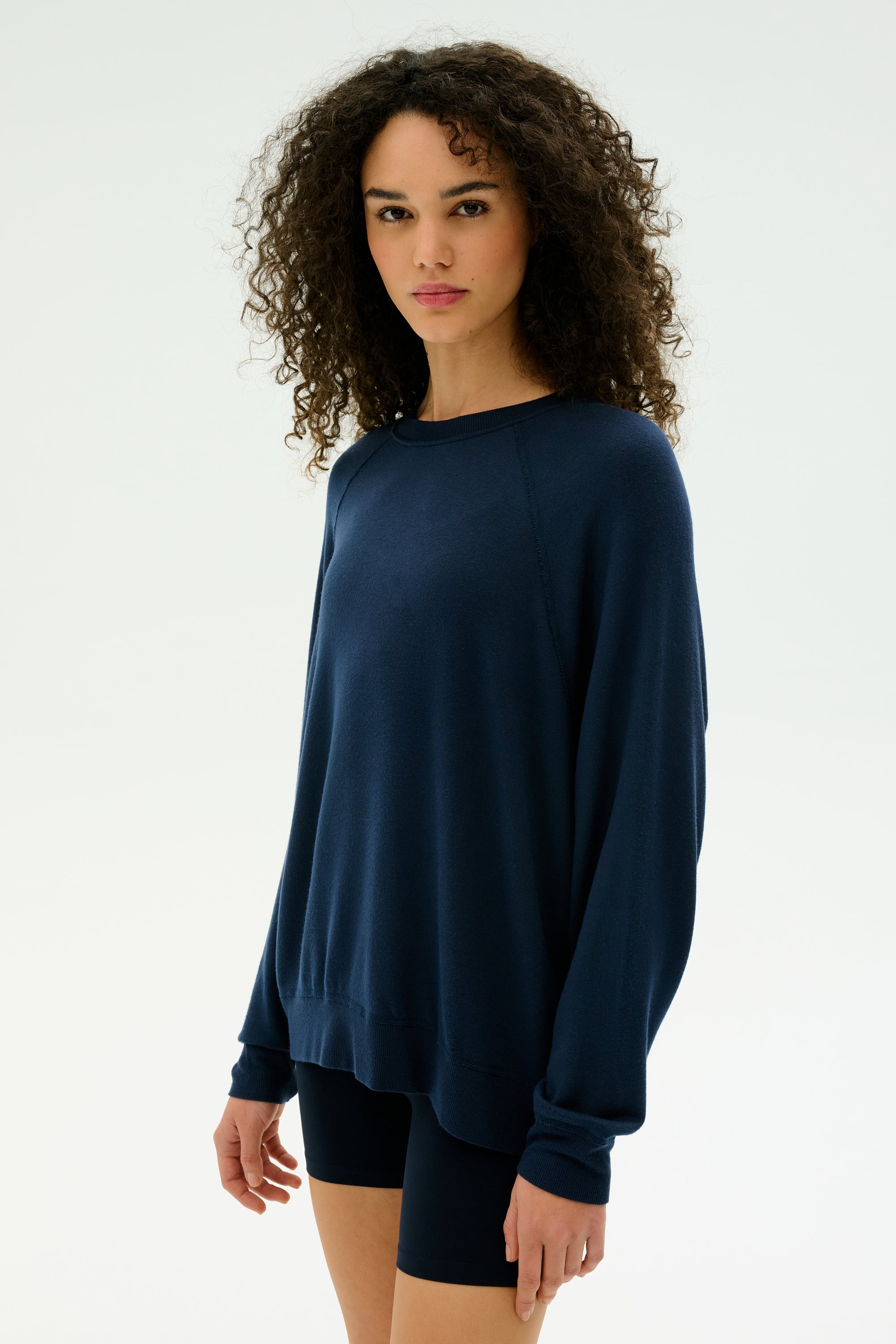 A person with curly hair wears the SPLITS59 Andie Fleece Sweatshirt in Indigo and black shorts, standing against a plain light background.