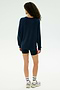 Someone with curly hair stands facing away on a plain light background, wearing the SPLITS59 Andie Fleece Sweatshirt in Indigo, paired with black shorts, white socks, and sneakers.