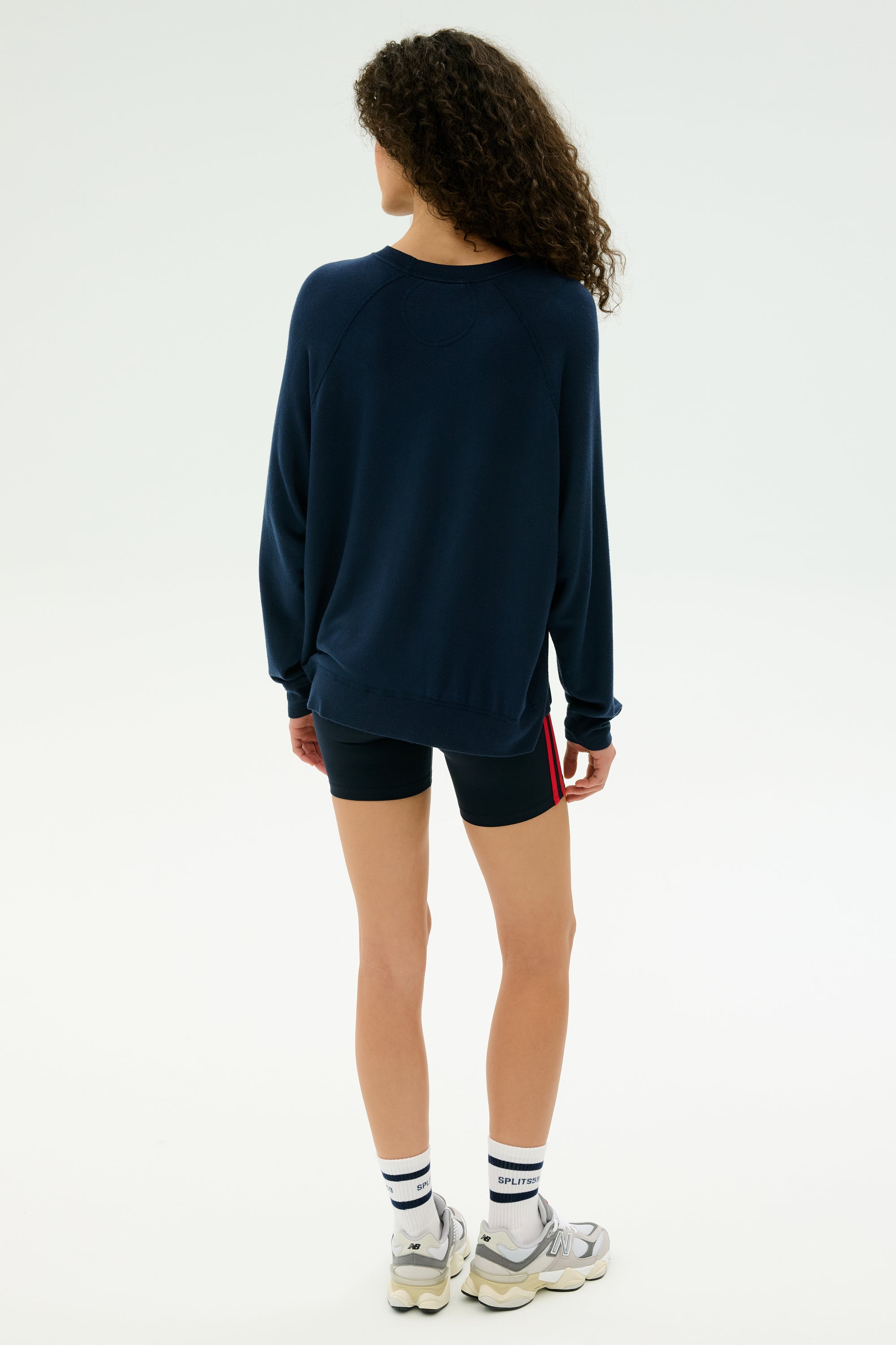 Someone with curly hair stands facing away on a plain light background, wearing the SPLITS59 Andie Fleece Sweatshirt in Indigo, paired with black shorts, white socks, and sneakers.