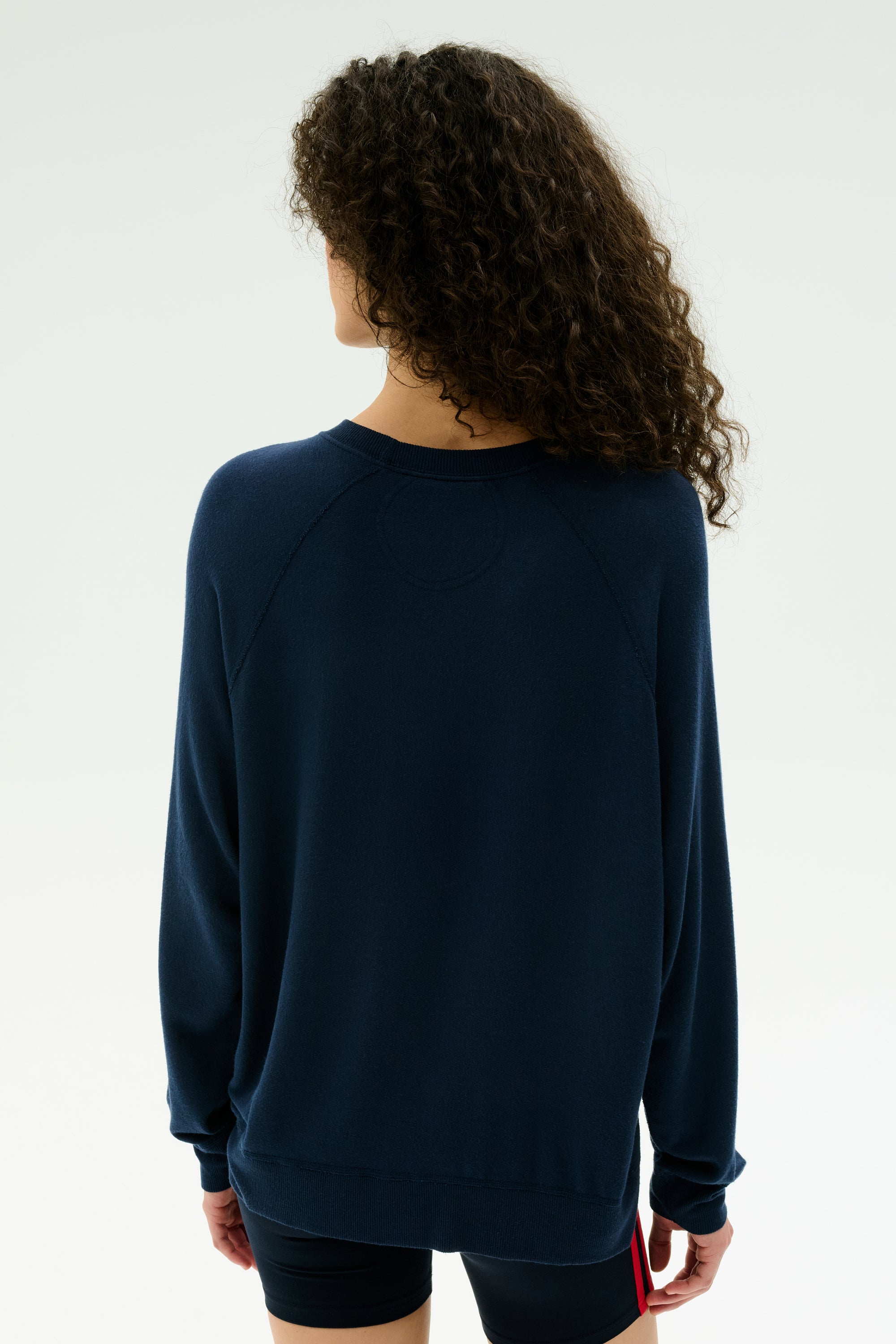A person with curly hair stands facing away, wearing the SPLITS59 Andie Fleece Sweatshirt in Indigo and black shorts against a plain, light background.