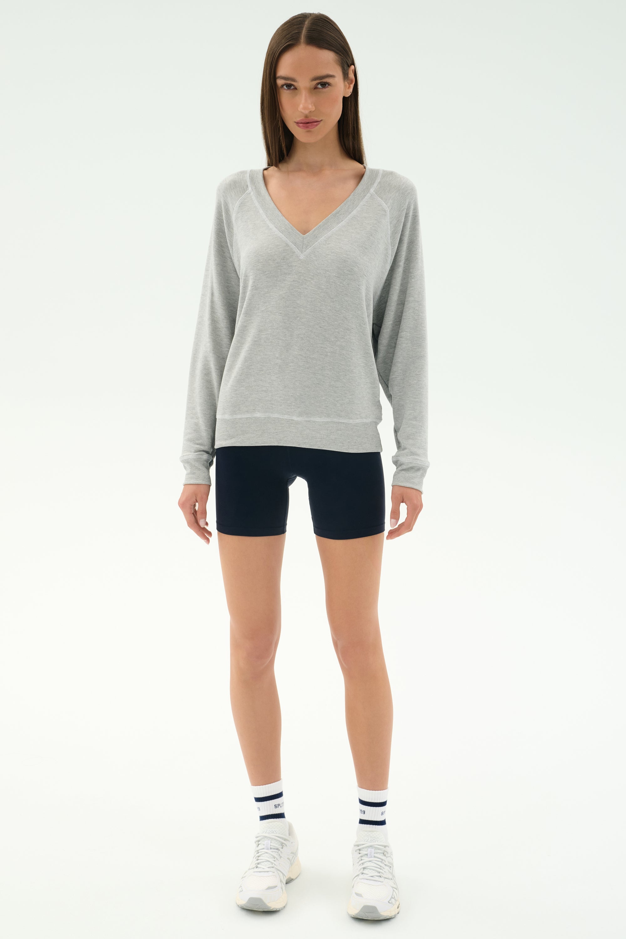 A woman faces forward against a white background wearing the SPLITS59 Bennie Fleece V Neck Sweatshirt in Heather Grey/White, paired with black shorts, white socks with black stripes, and white sneakers.