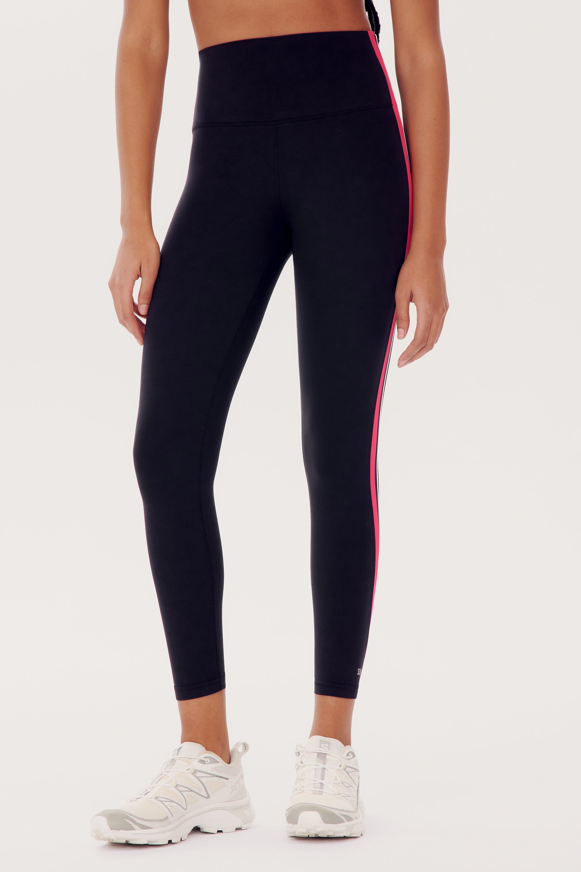 A person wearing SPLITS59 Bianca High Waist Rigor 7/8 - Black leggings with a red stripe along the side and white sneakers.