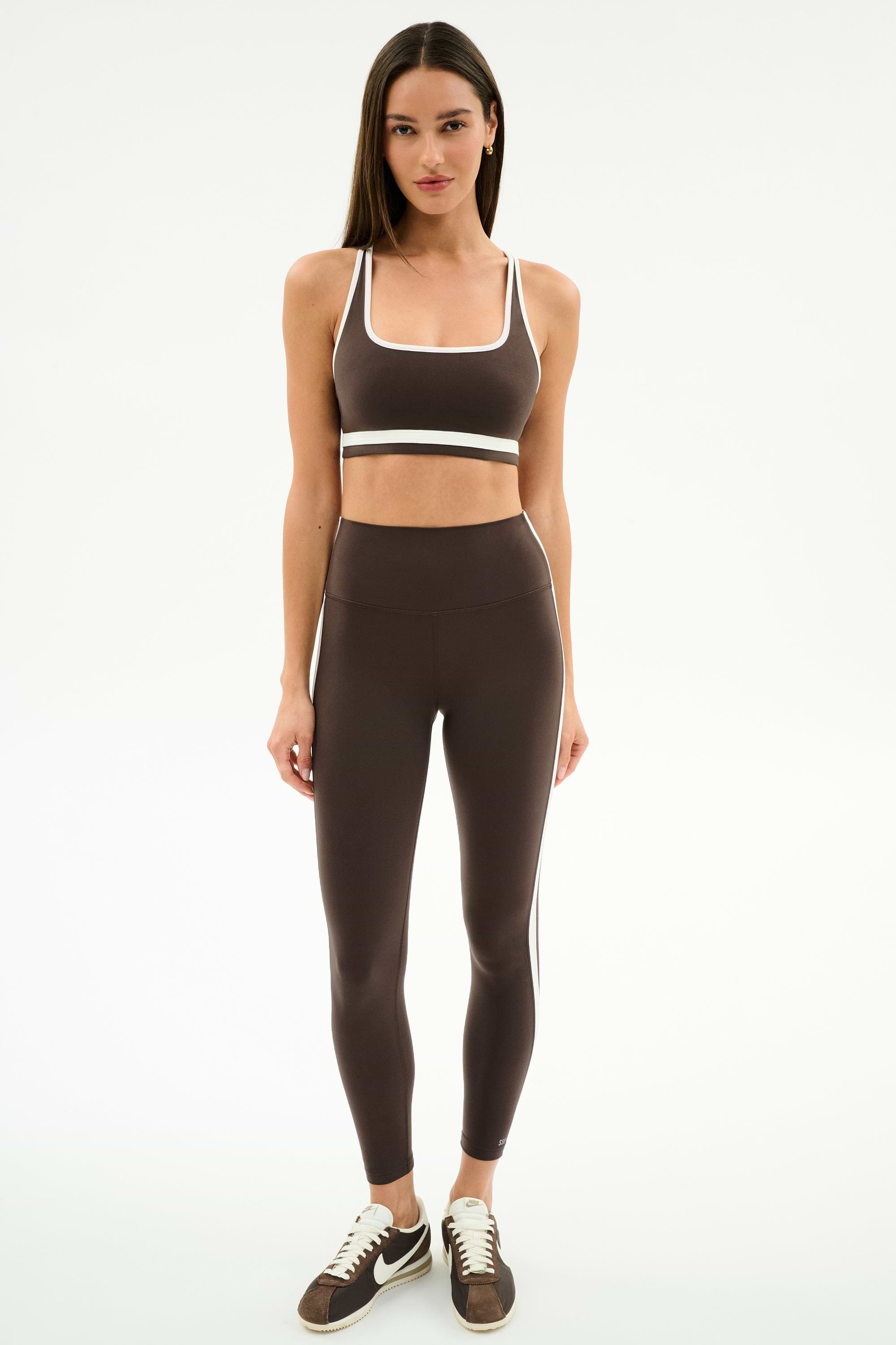 A woman faces forward wearing the SPLITS59 Bianca Rigor Bra in Dark Chocolate/White with matching high-waisted leggings and brown Nike sneakers—an ideal gym workout outfit.
