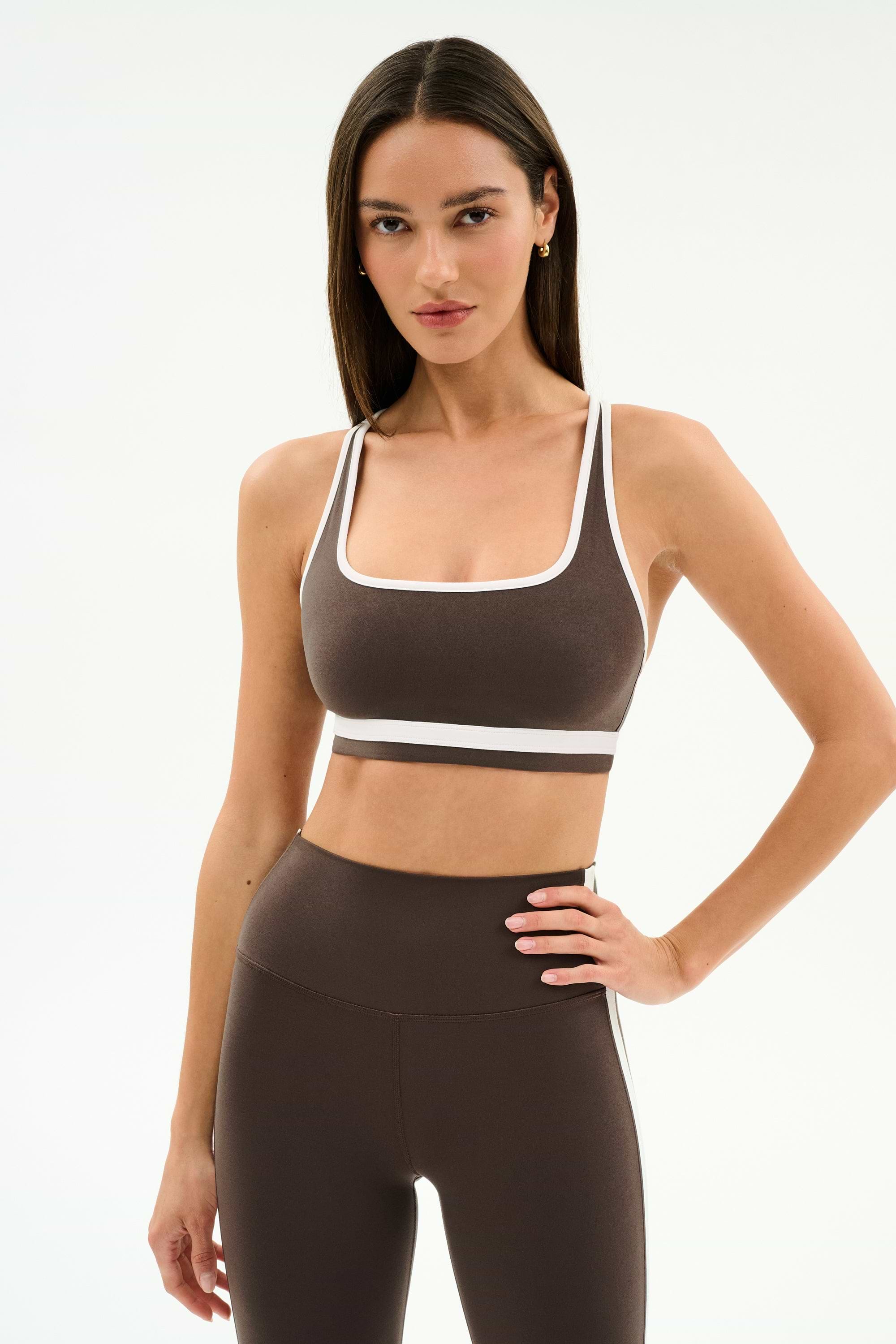 A woman stands against a plain white background wearing the SPLITS59 Bianca Rigor Bra in Dark Chocolate/White with matching high-waisted leggings, an ideal set for high-impact workouts.