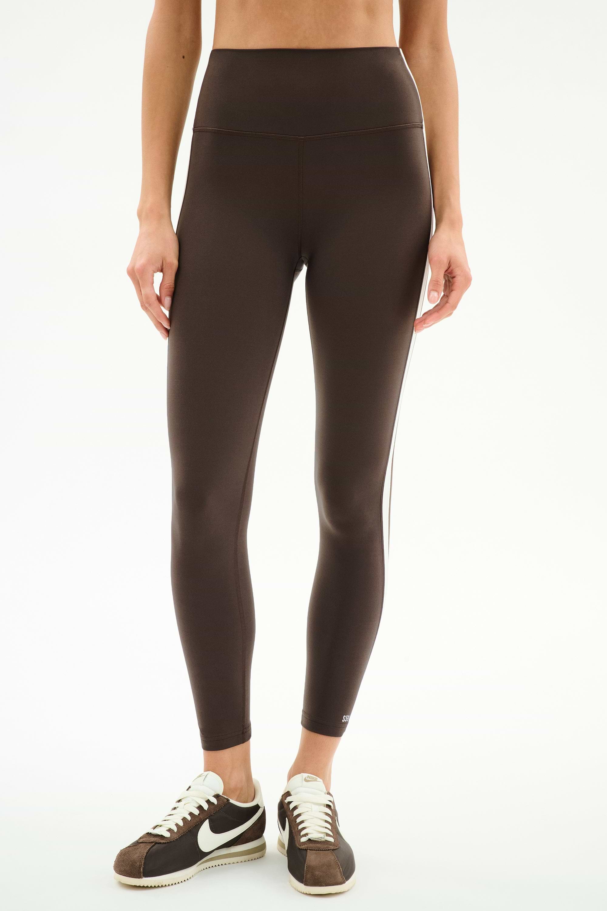 A person wears SPLITS59 Bianca High Waist Rigor 7/8 leggings in Dark Chocolate/White with brown-and-white sneakers, standing against a plain white background.