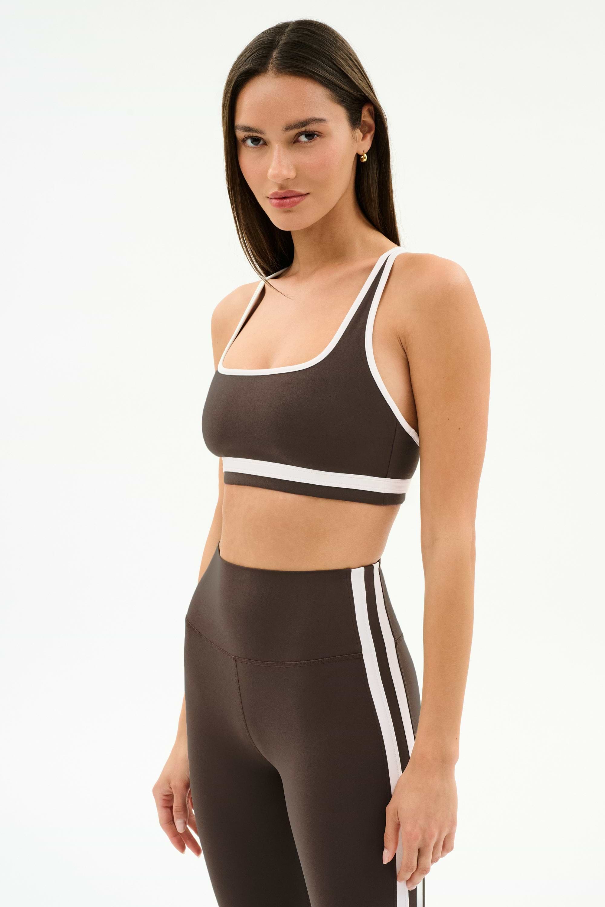A woman in a SPLITS59 Bianca Rigor Bra - Dark Chocolate/White and coordinating high-waisted leggings with white trim, ideal for gym workouts, stands against a plain white background.