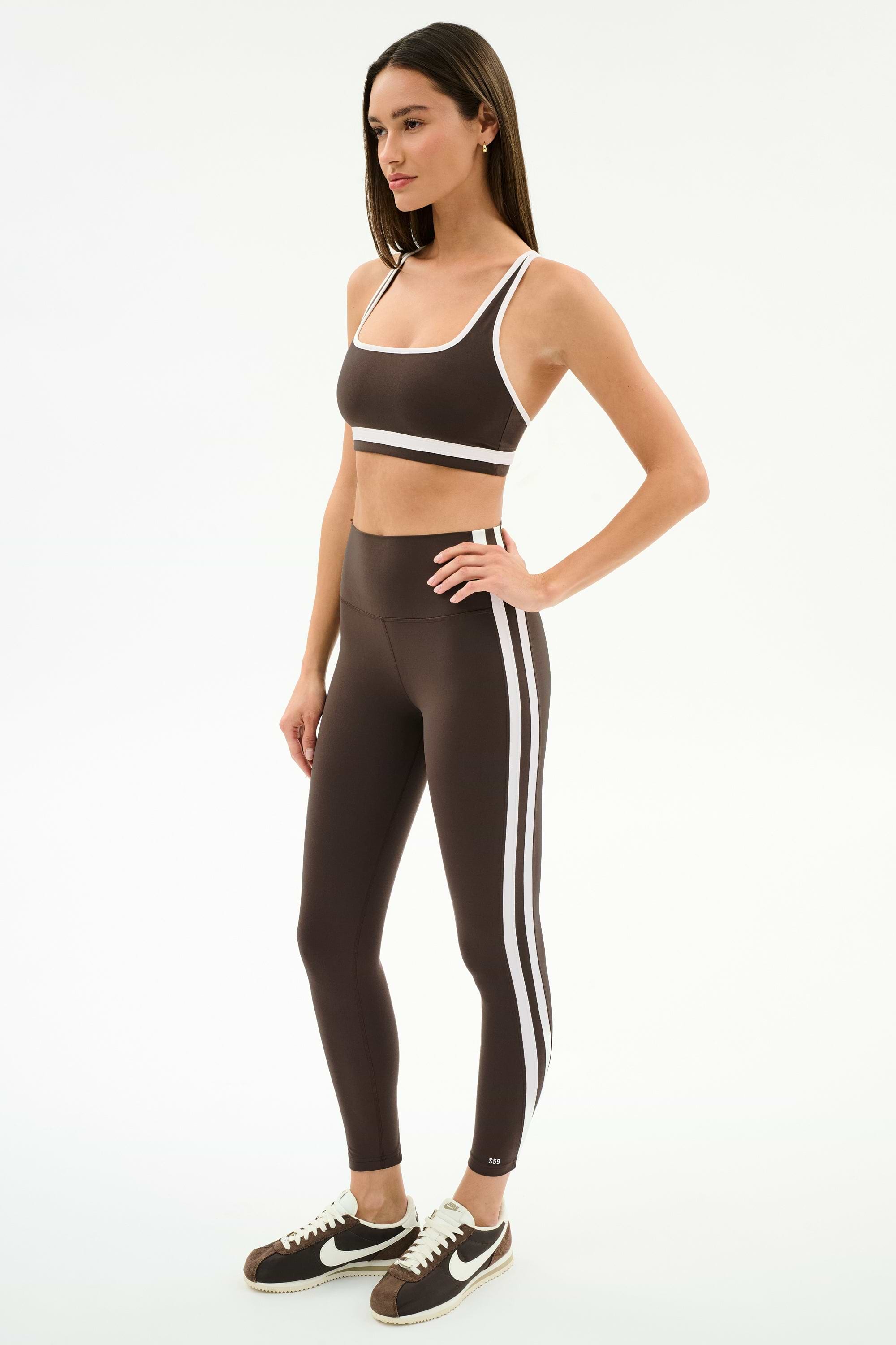 A woman wears the SPLITS59 Bianca Rigor Bra in Dark Chocolate/White and matching high-waisted leggings with white stripes, paired with black and white Nike sneakers—an ideal gym outfit—against a plain white background.