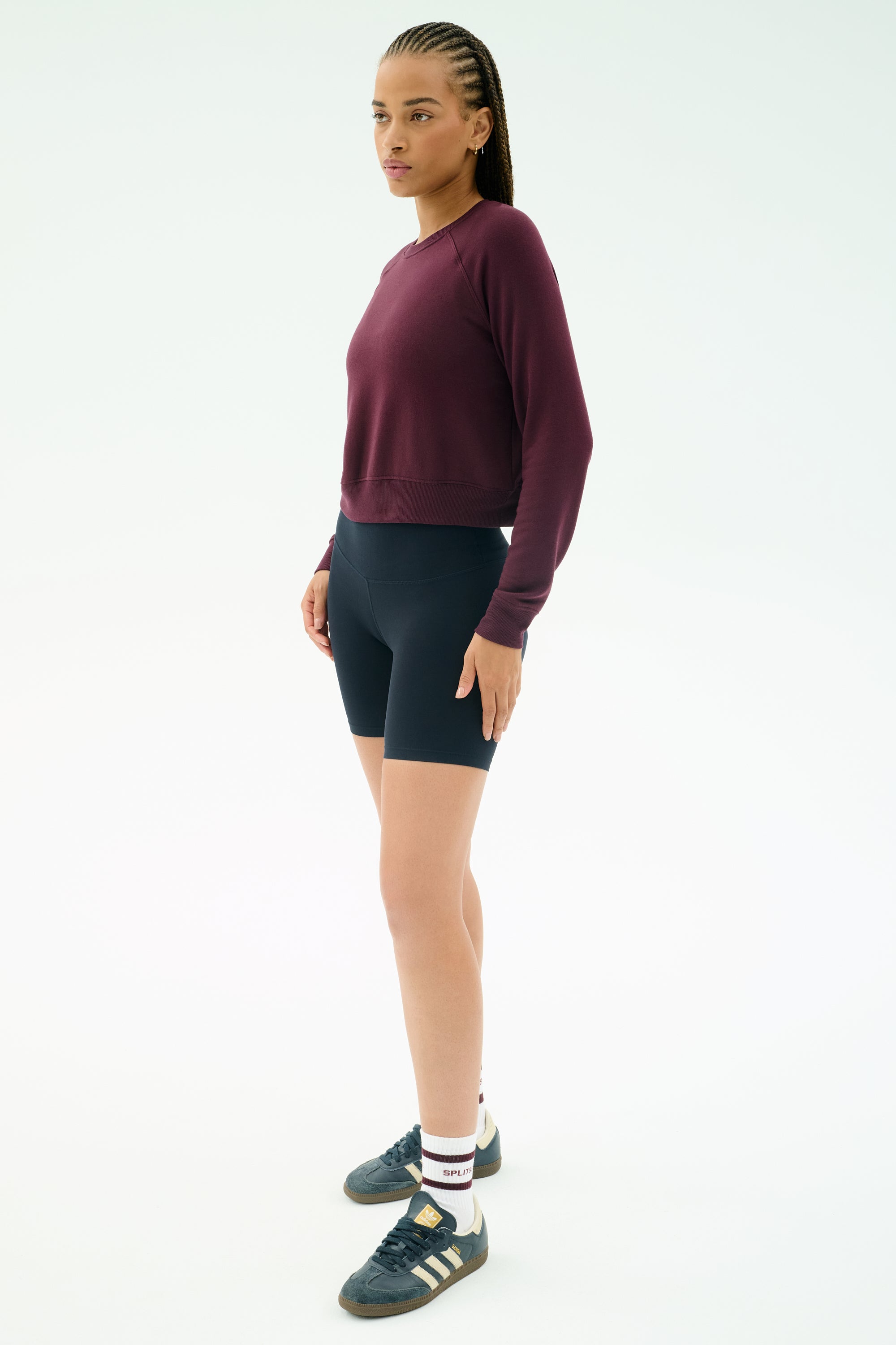 A person stands against a plain background wearing the SPLITS59 Brooks Fleece Long Sleeve Sweatshirt in Dark Cherry, paired with black bike shorts, white socks, and black Adidas sneakers.
