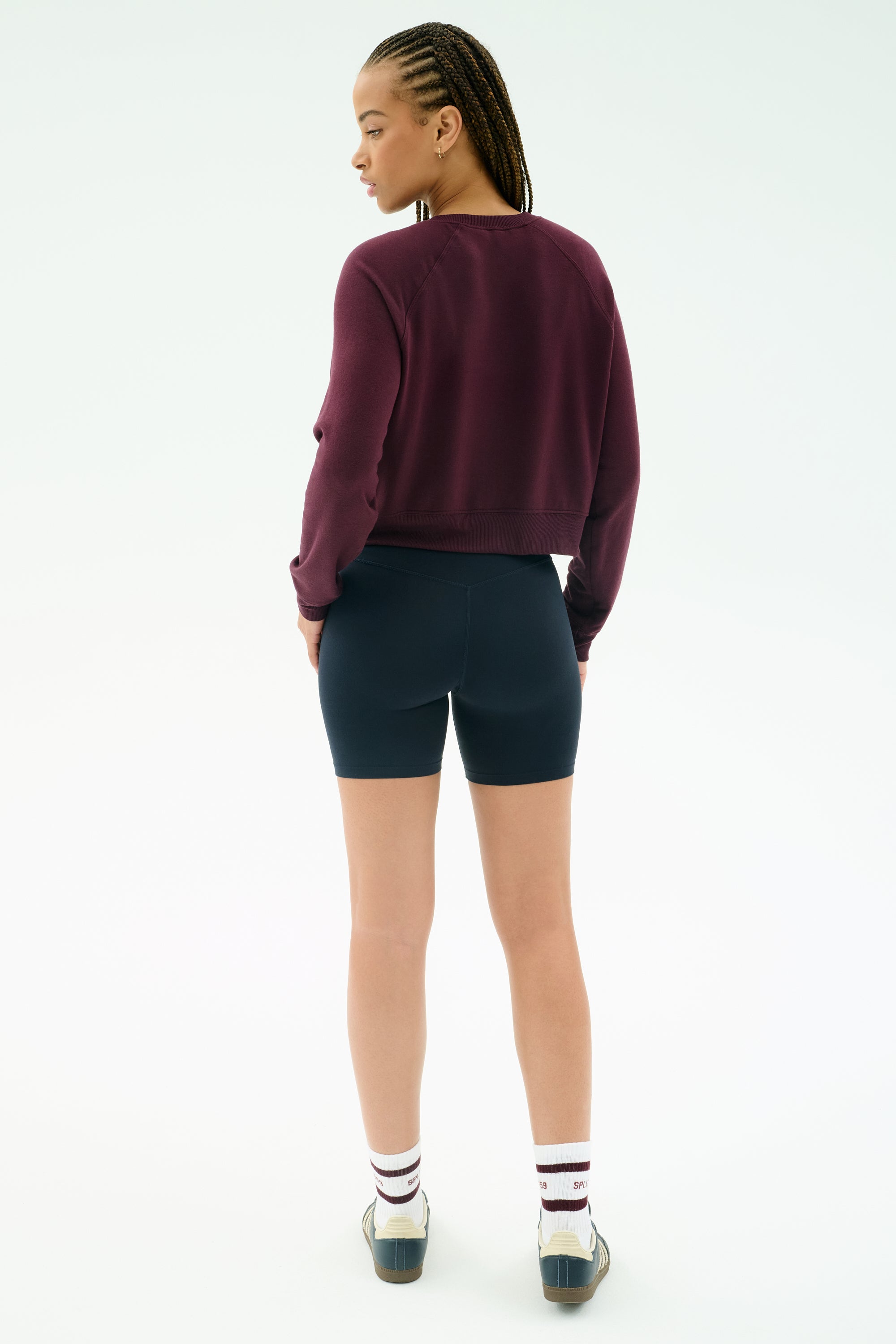 A person with braided hair wears a SPLITS59 Brooks Fleece Long Sleeve Sweatshirt in Dark Cherry, black shorts, white socks, and sneakers, standing with their back to a plain light background.