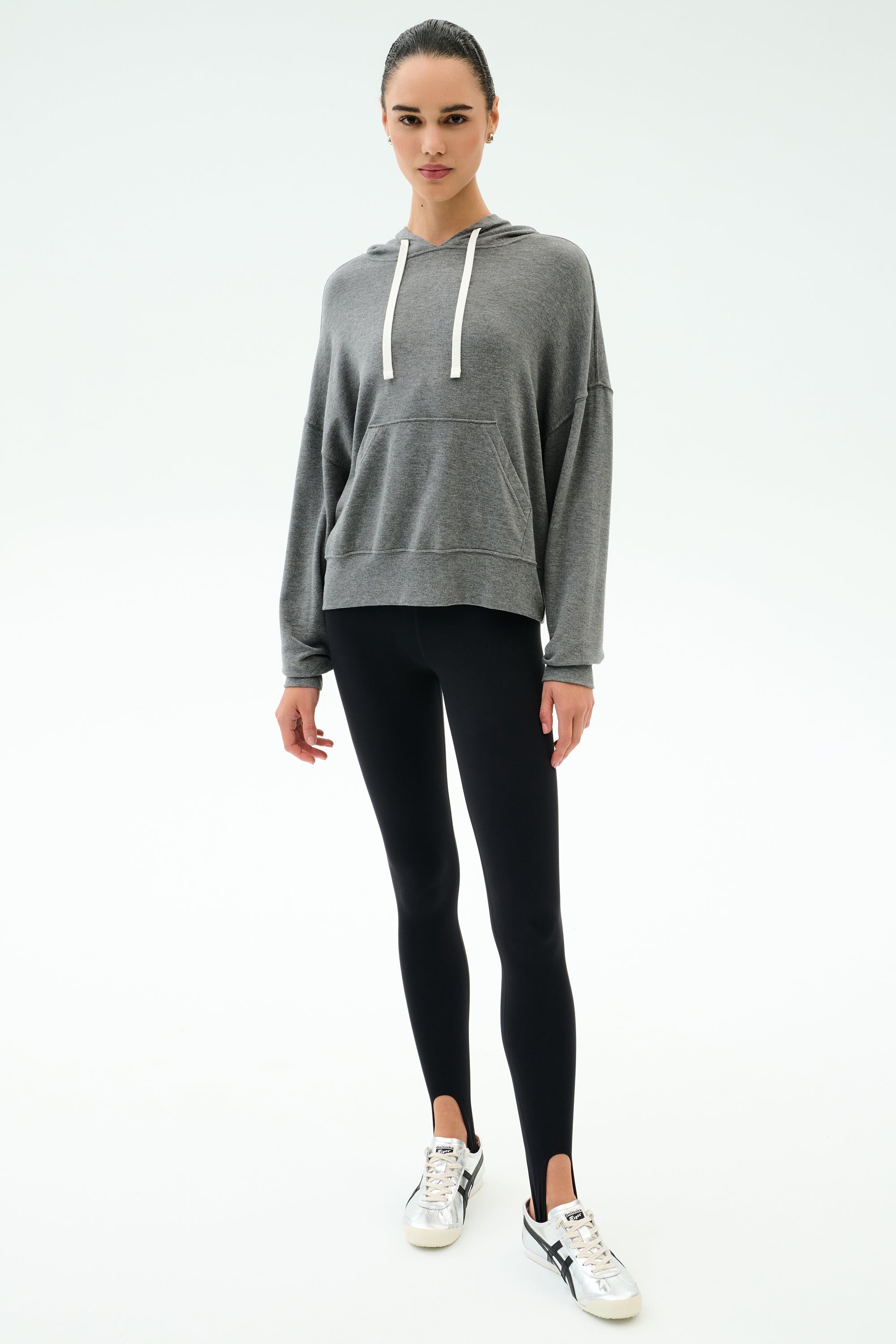 A person stands facing forward, wearing the SPLITS59 Joey Long Sleeve Hoodie in Medium Heather Grey, black spandex leggings, and white sneakers against a plain white background.