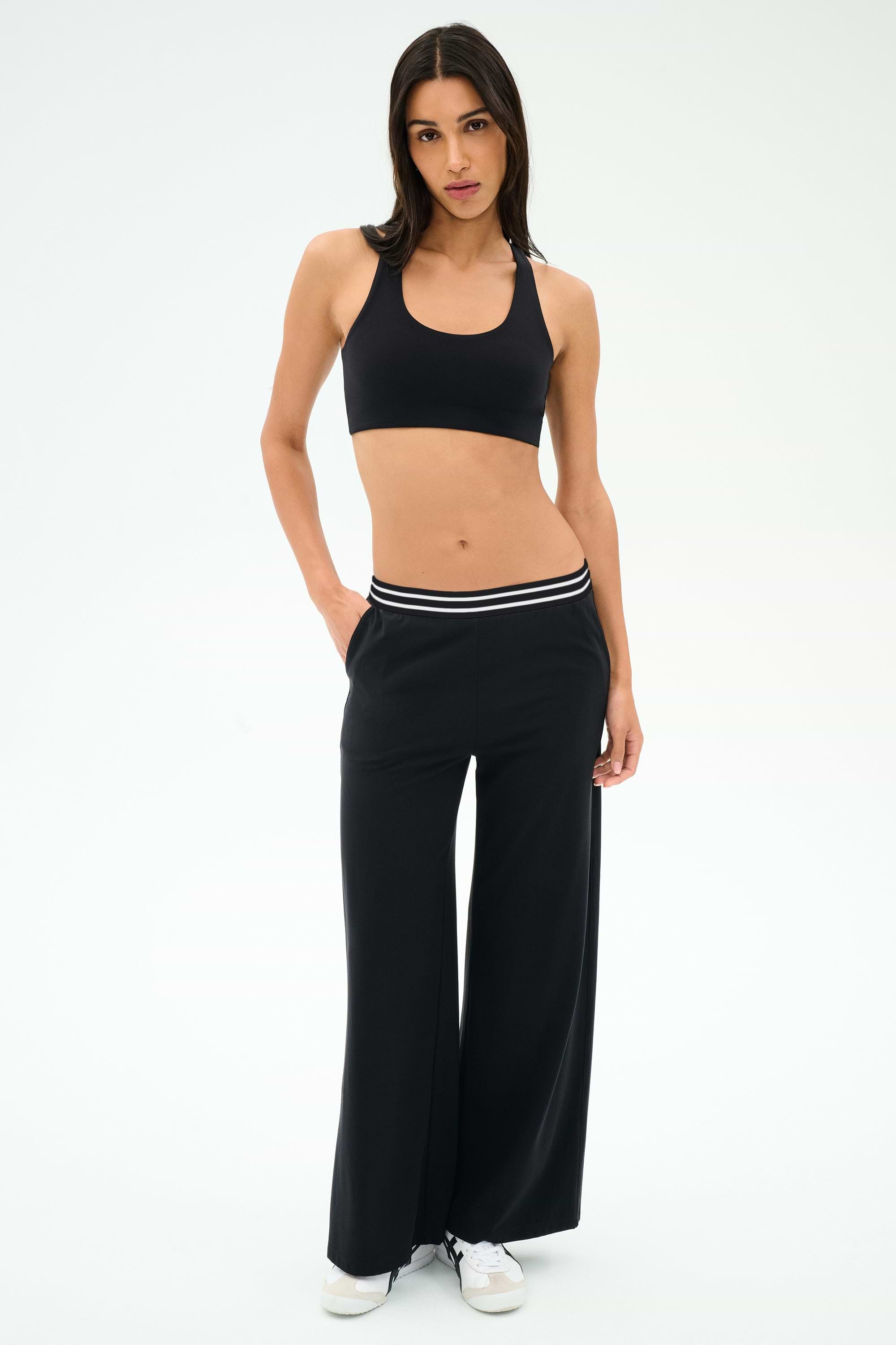 A woman wears the SPLITS59 Michele Airweight Trouser in Black with a white stripe waistband, paired with a black sports bra and white sneakers, standing against a plain white background.