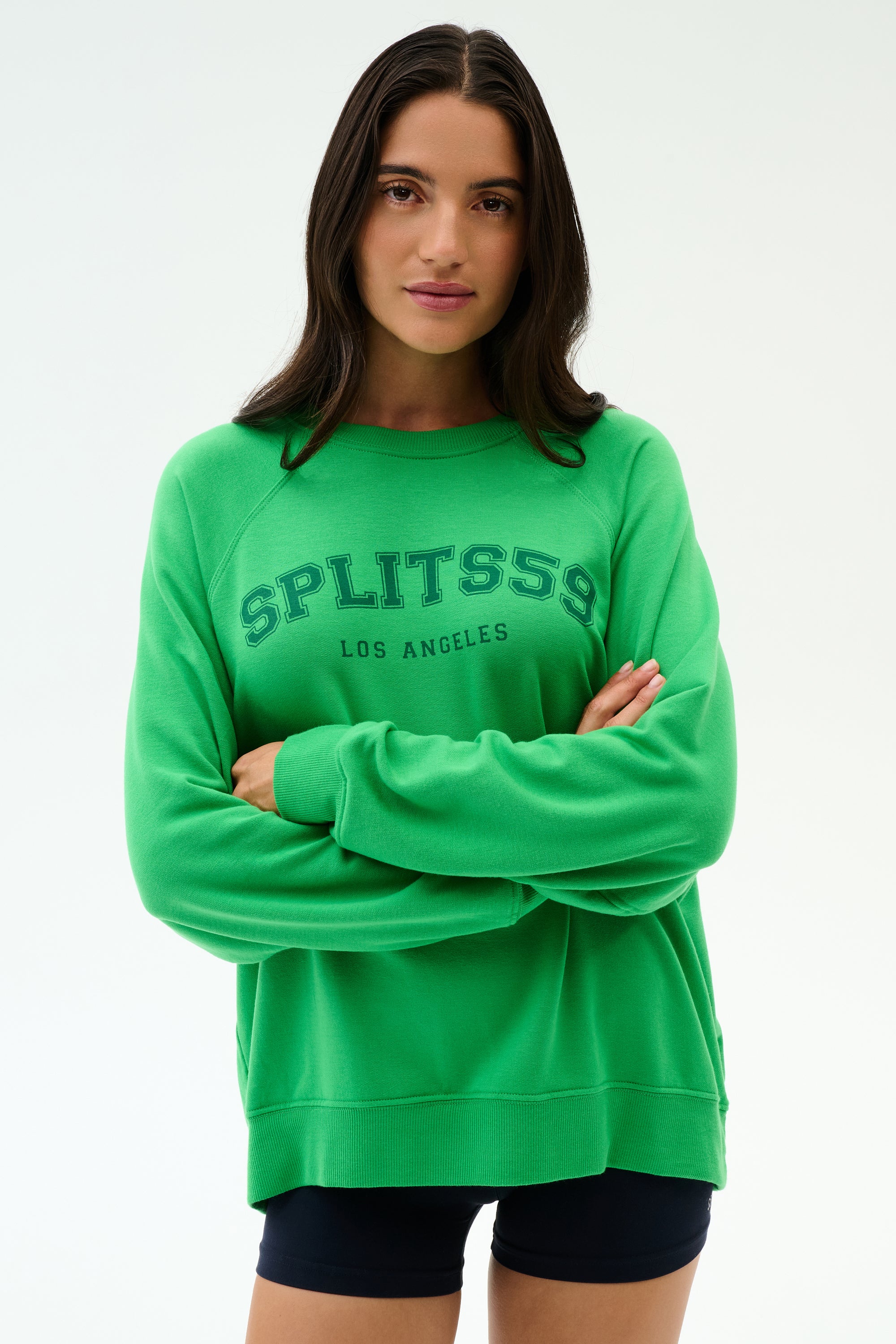 A woman with long dark hair wears the SPLITS59 Andie Oversized Fleece Varsity Graphic Sweatshirt in Grass and black shorts, standing against a plain white background with arms crossed.