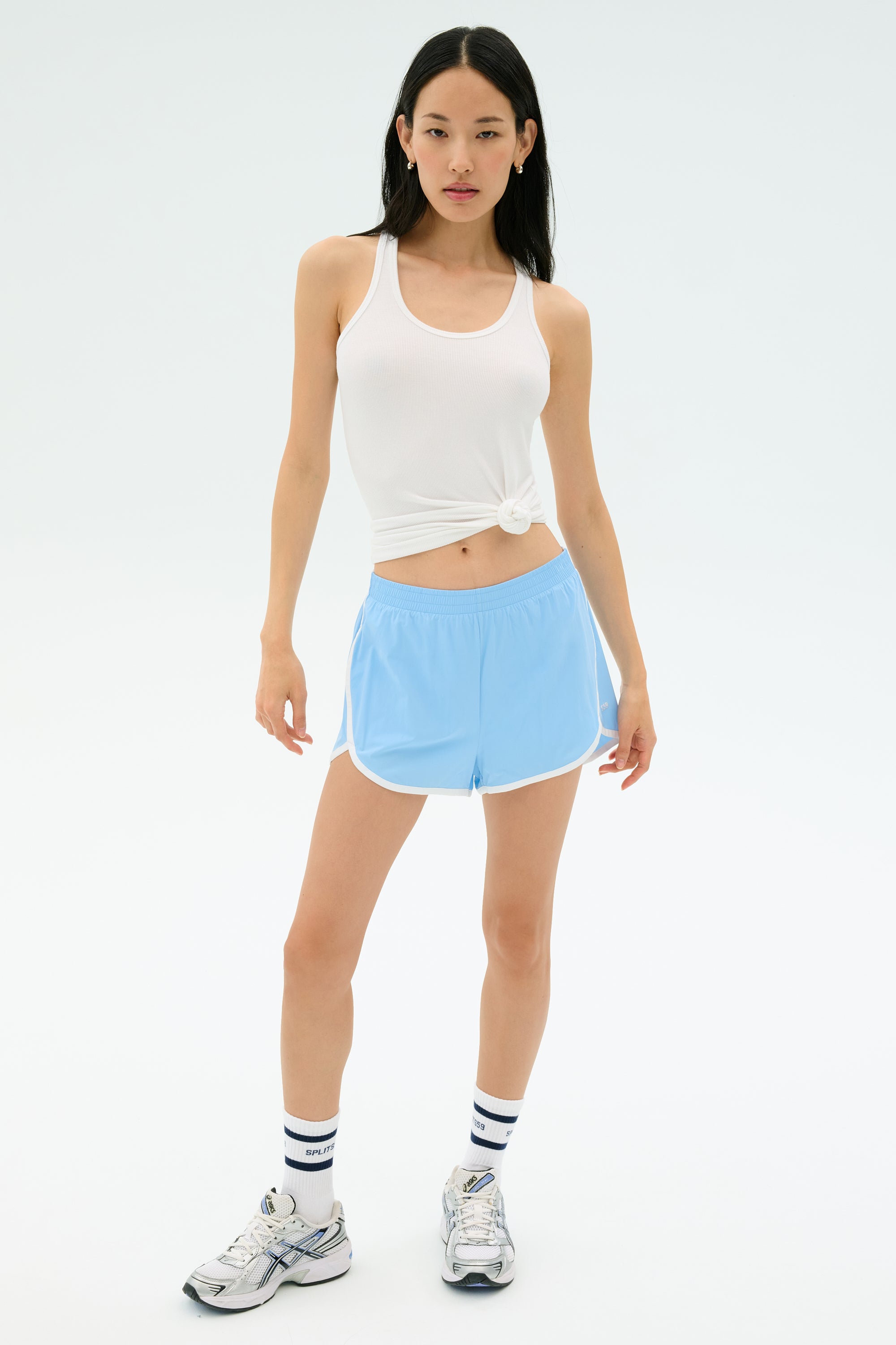 A woman stands against a plain background wearing the SPLITS59 Megan Woven Nylon Run Short in Skylight, a white tank top, striped socks, and athletic shoes.