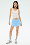 A woman stands against a plain background wearing the SPLITS59 Megan Woven Nylon Run Short in Skylight, a white tank top, striped socks, and athletic shoes.