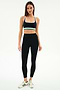 A woman stands facing forward in SPLITS59 athletic wear: a black sports bra with white and green accents, Bianca High Waist Rigor 7/8 leggings in Black/White, and white sneakers with green stripes.