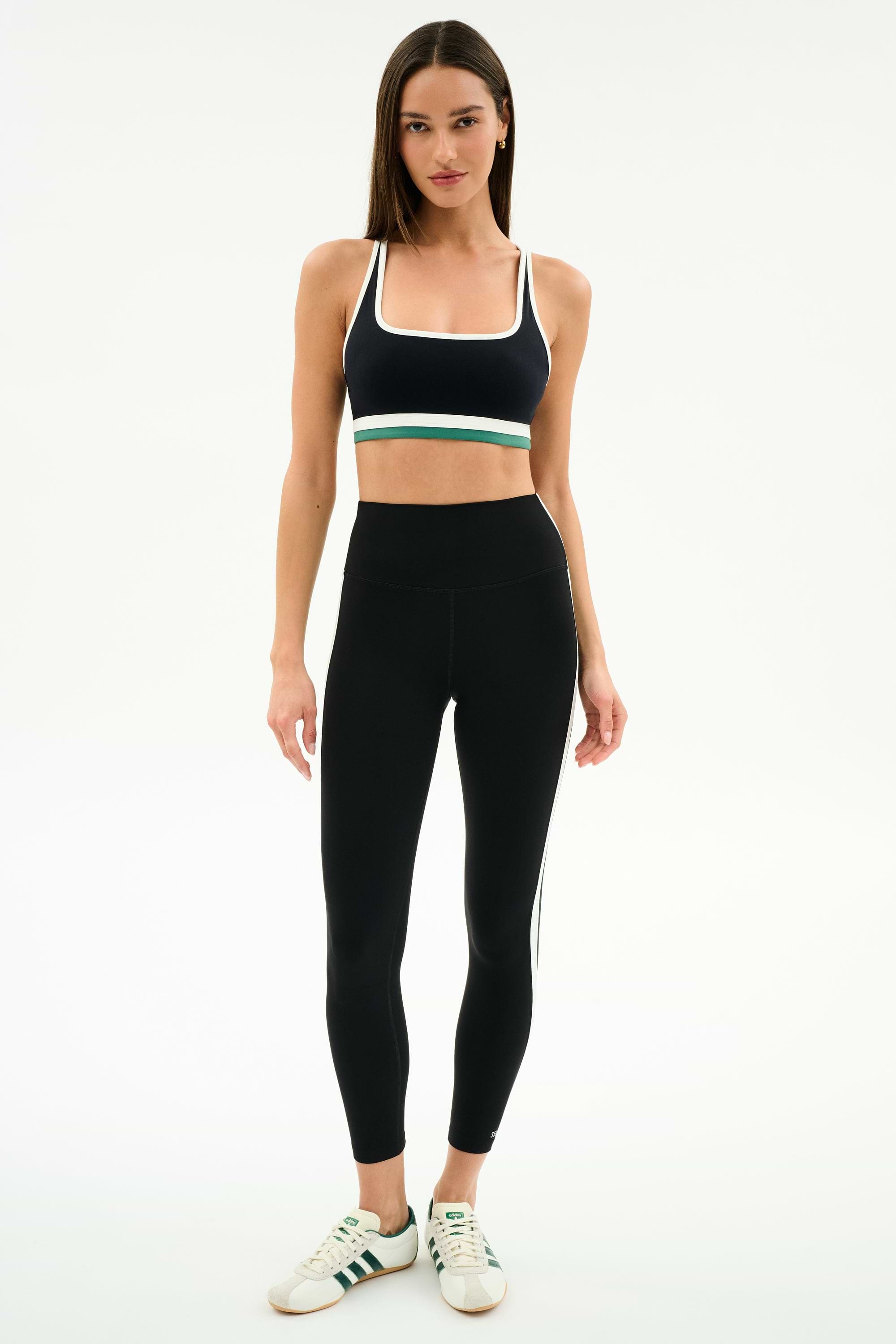 A woman stands facing forward in SPLITS59 athletic wear: a black sports bra with white and green accents, Bianca High Waist Rigor 7/8 leggings in Black/White, and white sneakers with green stripes.