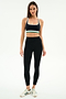 A woman stands facing forward in SPLITS59 athletic wear: a black sports bra with white and green accents, Bianca High Waist Rigor 7/8 leggings in Black/White, and white sneakers with green stripes.