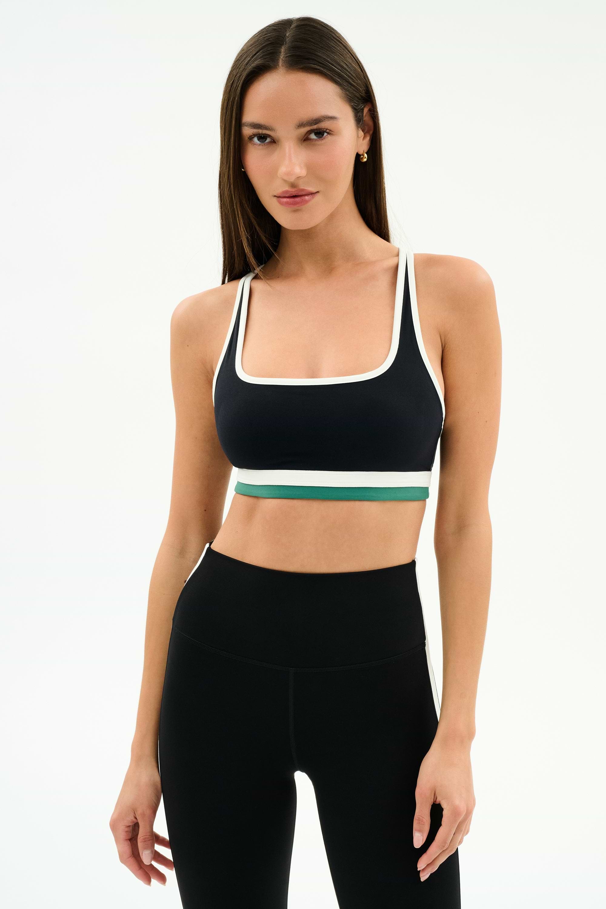 A woman stands against a plain background wearing the SPLITS59 Bianca Rigor Bra in Black/White and high-waisted black spandex leggings.