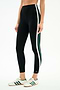 A person wears SPLITS59's Bianca High Waist Rigor 7/8 leggings in Black/White, featuring white and green side stripes, paired with white sneakers with green stripes, standing against a plain background.