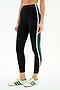 A person wears SPLITS59's Bianca High Waist Rigor 7/8 leggings in Black/White, featuring white and green side stripes, paired with white sneakers with green stripes, standing against a plain background.