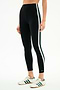 A person stands against a plain background in SPLITS59's Bianca High Waist Rigor 7/8 leggings in black/white, featuring white and green side stripes, paired with white Adidas sneakers accented by green stripes.