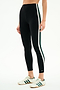 A person stands against a plain background in SPLITS59's Bianca High Waist Rigor 7/8 leggings in black/white, featuring white and green side stripes, paired with white Adidas sneakers accented by green stripes.