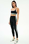 A woman stands in a studio wearing the SPLITS59 Bianca High Waist Rigor 7/8 leggings in black/white—a matching black athletic set with green and white stripes—and white Adidas sneakers with green stripes.
