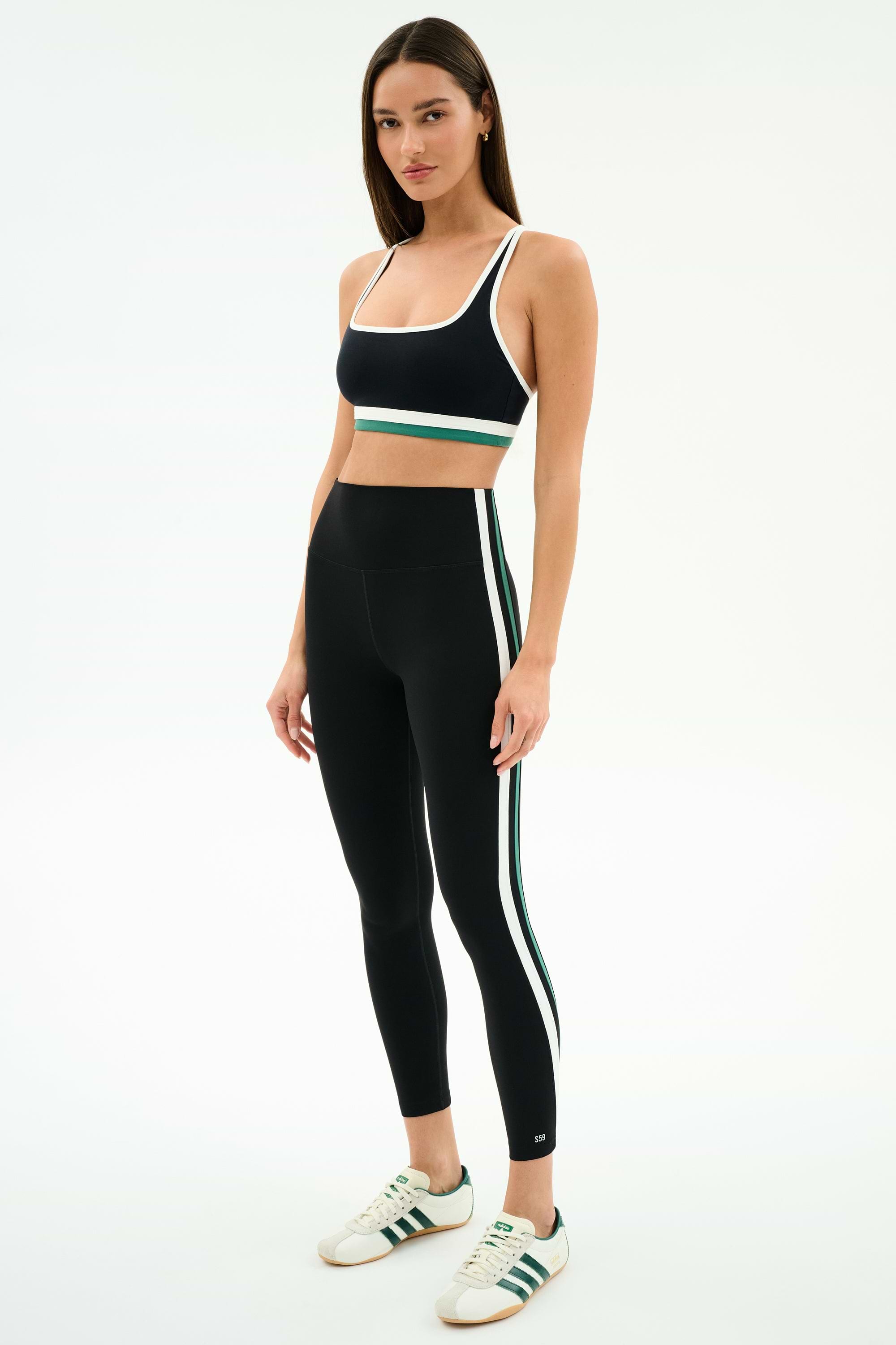 A woman stands in a studio wearing the SPLITS59 Bianca High Waist Rigor 7/8 leggings in black/white—a matching black athletic set with green and white stripes—and white Adidas sneakers with green stripes.