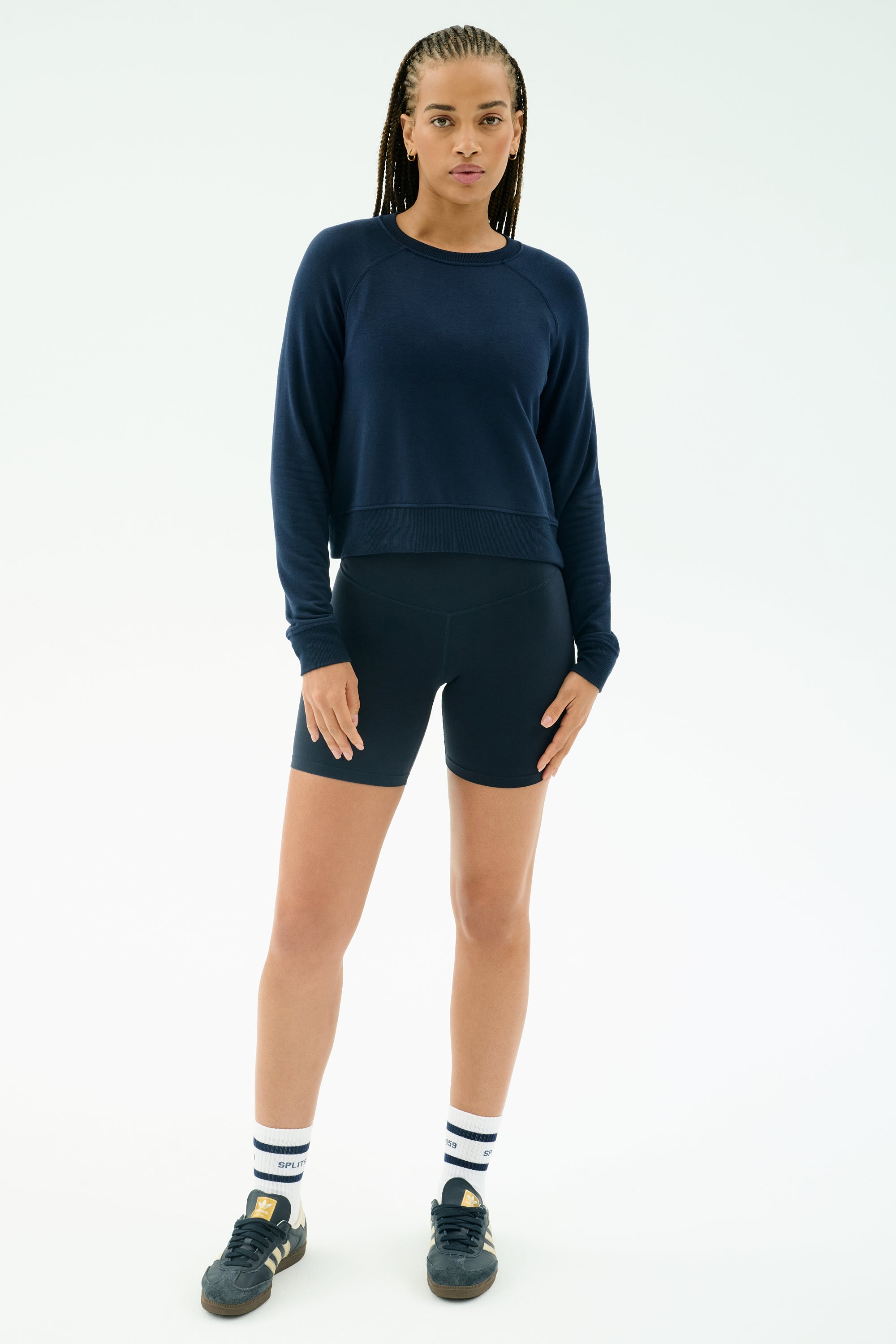 A person stands facing forward wearing the SPLITS59 Brooks Fleece Long Sleeve Sweatshirt in Indigo, navy shorts, striped white socks, and black sneakers against a plain white background.