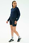 A person stands against a plain white background wearing the SPLITS59 Brooks Fleece Long Sleeve Sweatshirt in indigo, navy bike shorts, white socks, and navy sneakers.