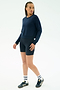 A person stands against a plain white background wearing the SPLITS59 Brooks Fleece Long Sleeve Sweatshirt in indigo, navy bike shorts, white socks, and navy sneakers.