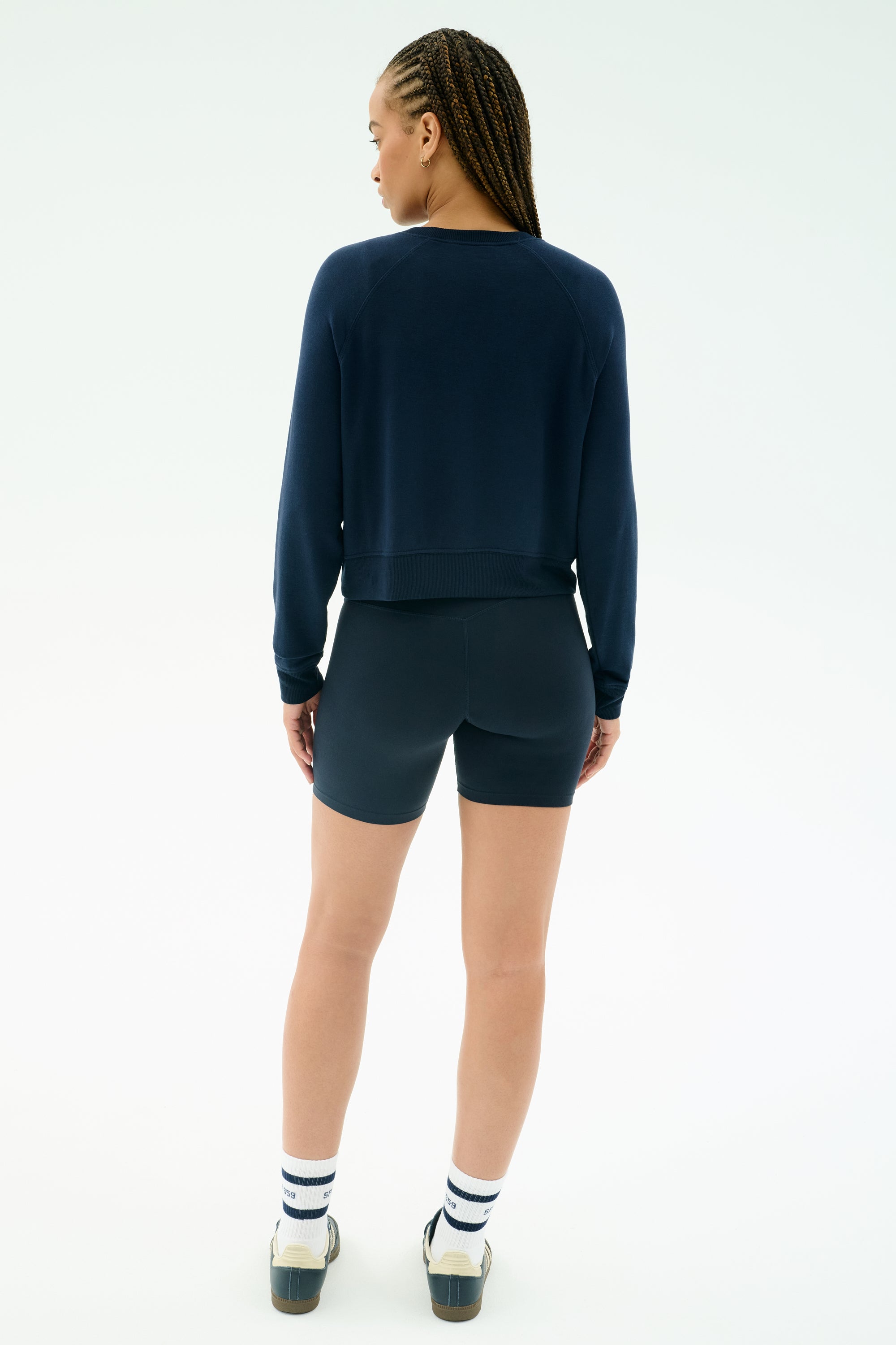 A person stands facing away, wearing the SPLITS59 Brooks Fleece Long Sleeve Sweatshirt in indigo, navy cycling shorts, white socks with blue stripes, and white sneakers against a plain light background.