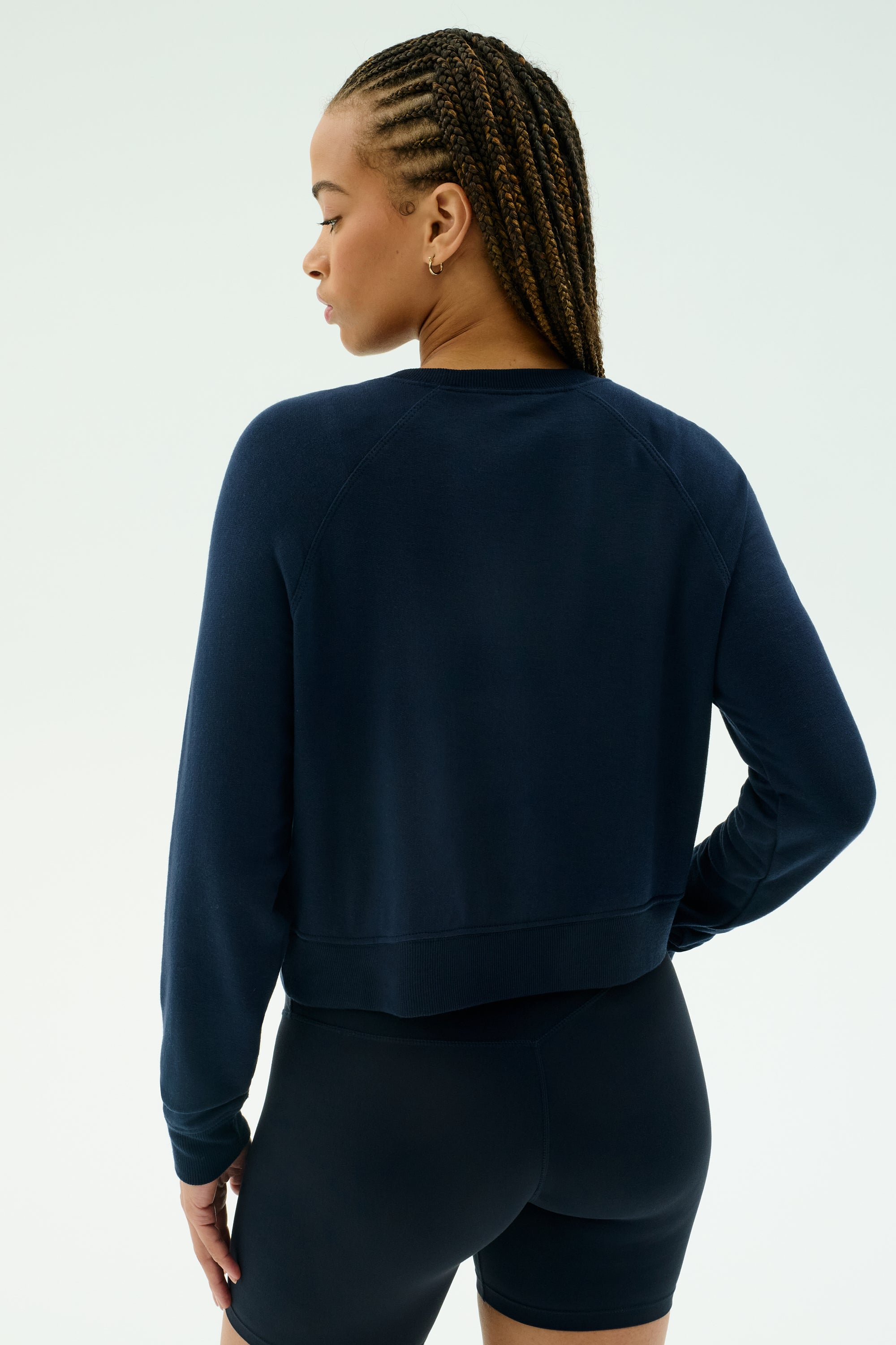A person with braided hair wears the SPLITS59 Brooks Fleece Long Sleeve Sweatshirt in Indigo and black shorts, standing facing away from the camera against a plain light background.