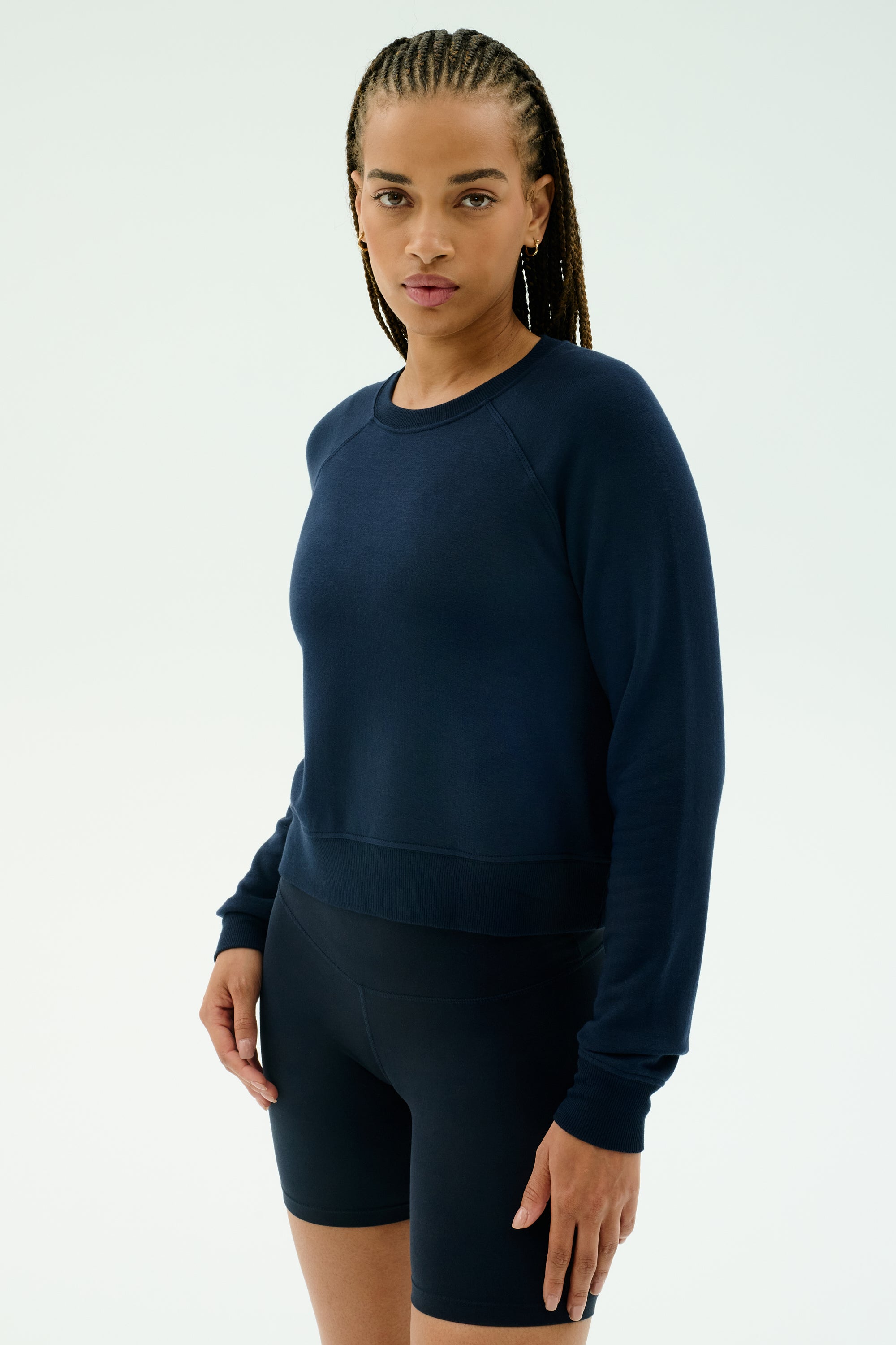 A woman with braided hair models the SPLITS59 Brooks Fleece Long Sleeve Sweatshirt in indigo and black biking shorts against a plain white background. Made in Los Angeles.