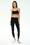 A woman faces forward wearing the SPLITS59 Eva Rigor Double Strap Bra in Black/Military, paired with matching black leggings featuring white stripes, and white sneakers with green accents.