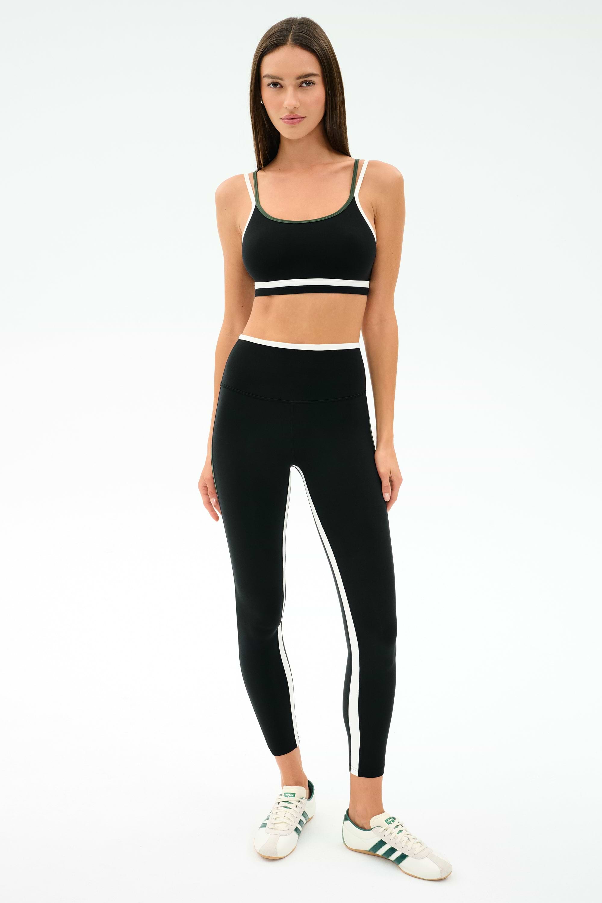 A woman stands facing forward, wearing the SPLITS59 Sam High Waist Rigor 7/8 leggings in Black/Military and a matching sports bra with white stripes, paired with white sneakers featuring green accents.